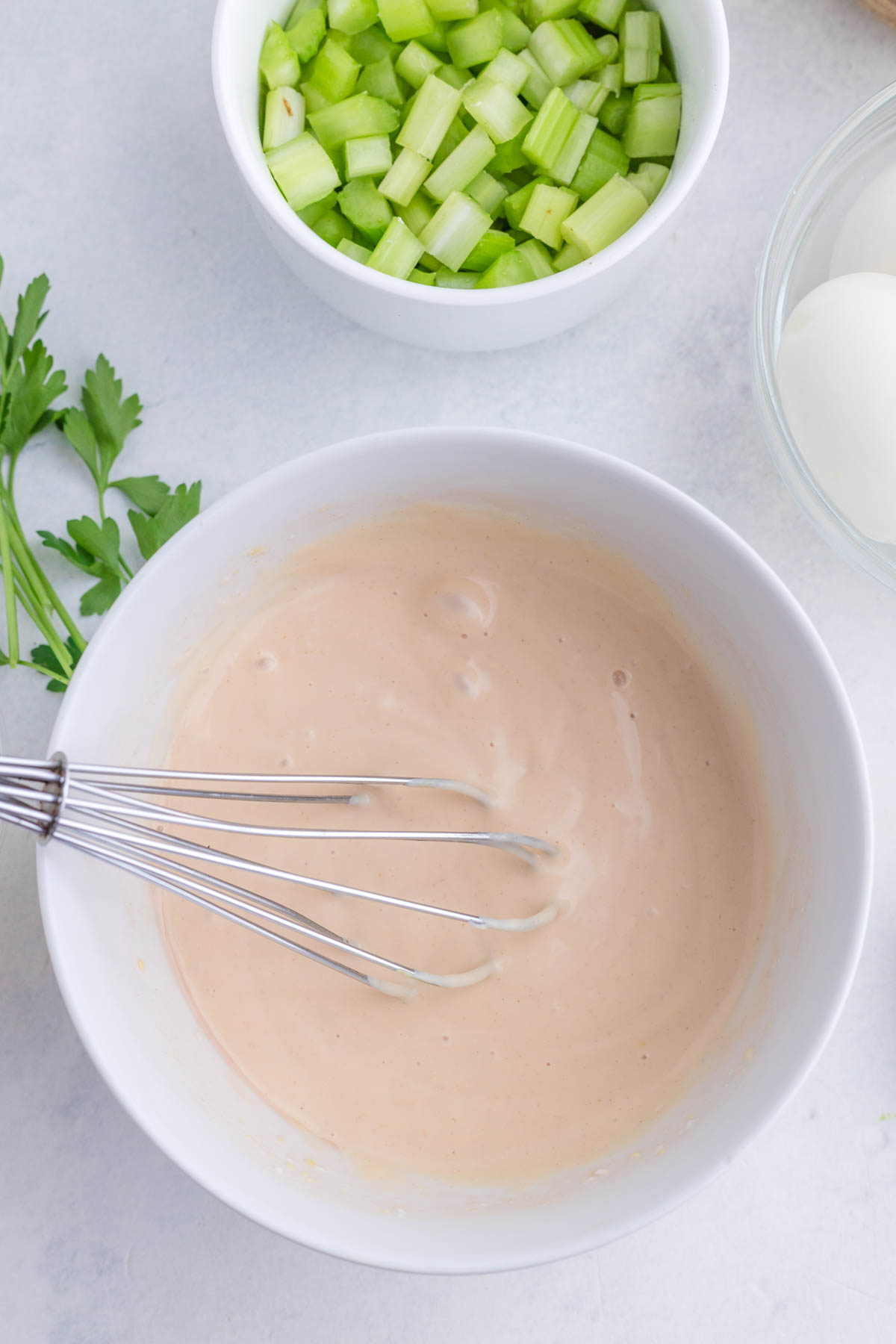 A bowl of creamy dressing with a whisk, next to bowls of chopped celery, hard-boiled eggs, and a sprig of parsley.