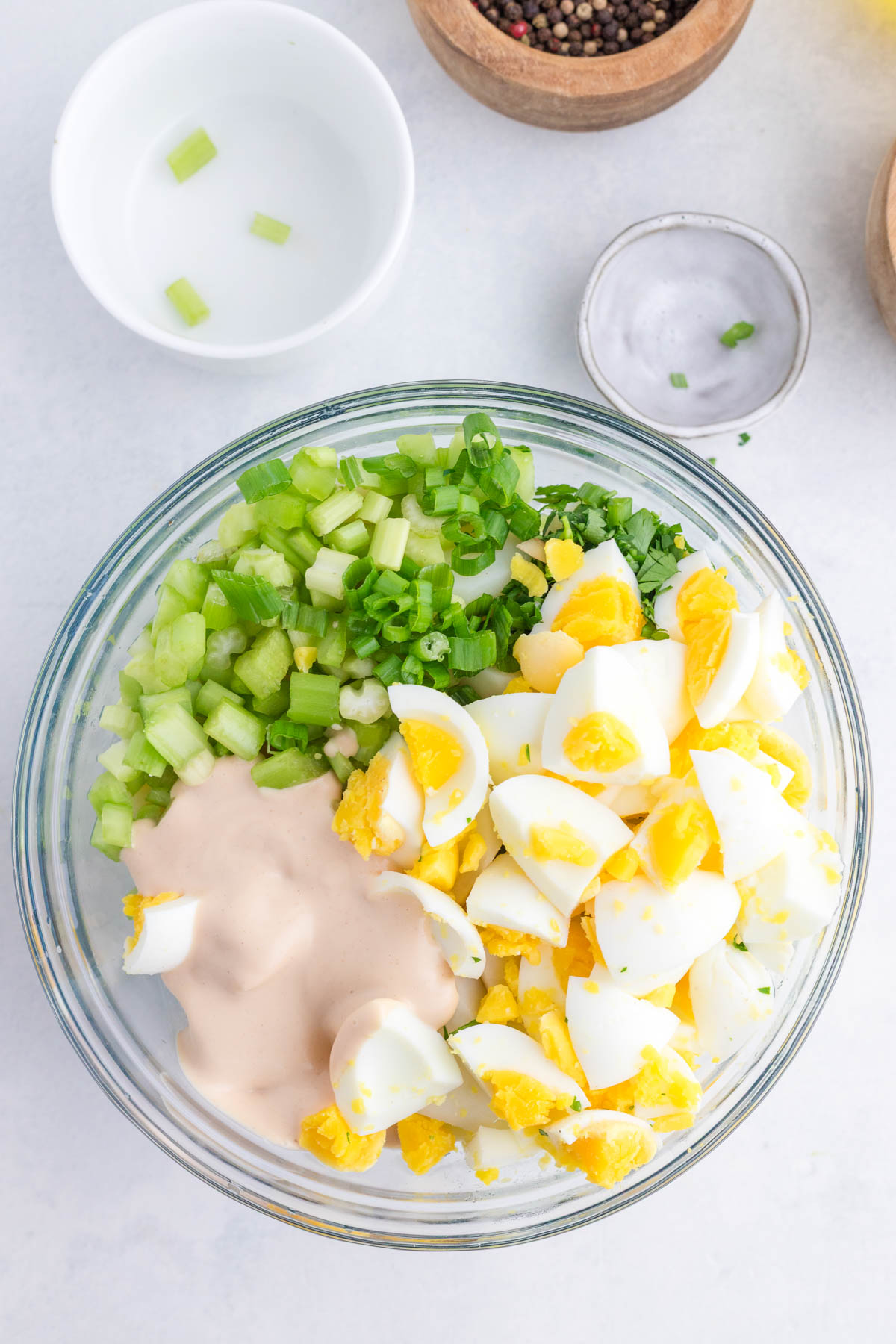 A glass bowl containing chopped hard-boiled eggs, celery, green onions, and salad dressing, on a white surface with small dishes nearby.