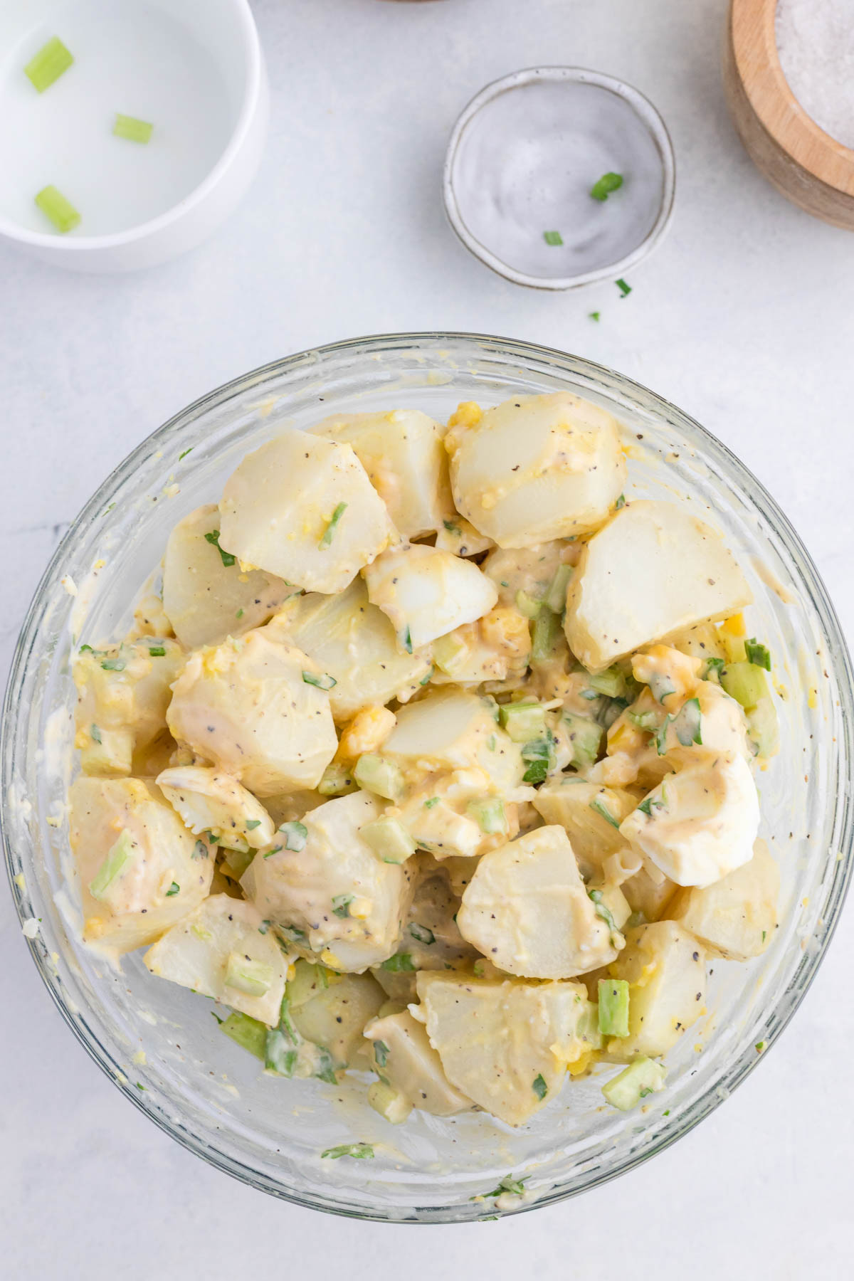 A glass bowl filled with potato salad mixed with chopped celery and herbs, with a small dish of salt and an empty bowl nearby on a white surface.