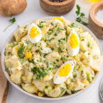 A bowl of potato salad with chopped celery and parsley, topped with quartered hard-boiled eggs, black pepper, and surrounded by bowls of salt and pepper.
