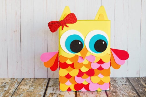 A colorful, handmade owl decoration made from paper with large blue eyes, a red bow, and layered feathers, standing on a wooden floor against a white plank wall.