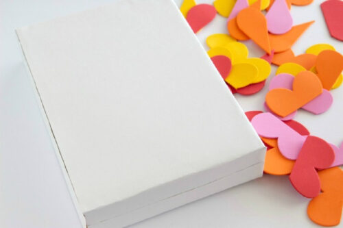 A closed plain white box sits next to a scattered pile of red, orange, and yellow paper hearts on a white surface.