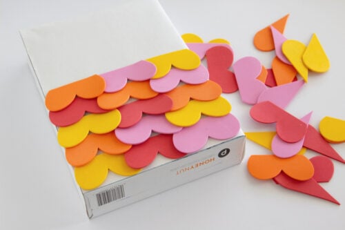 A box is partially covered with overlapping foam heart shapes in pink, orange, and yellow, with more foam shapes scattered nearby.