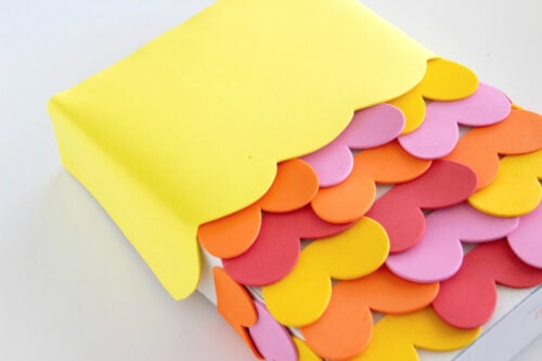 A rectangular object covered with overlapping heart-shaped pieces of foam in yellow, orange, pink, and red, with a yellow foam sheet partially wrapping the top.