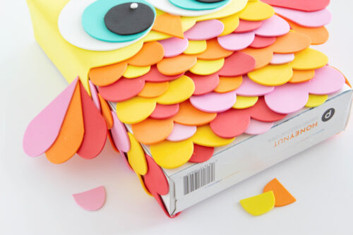 A rectangular box decorated as an owl with colorful foam cutouts for eyes, wings, and feathers in yellow, orange, pink, and red.