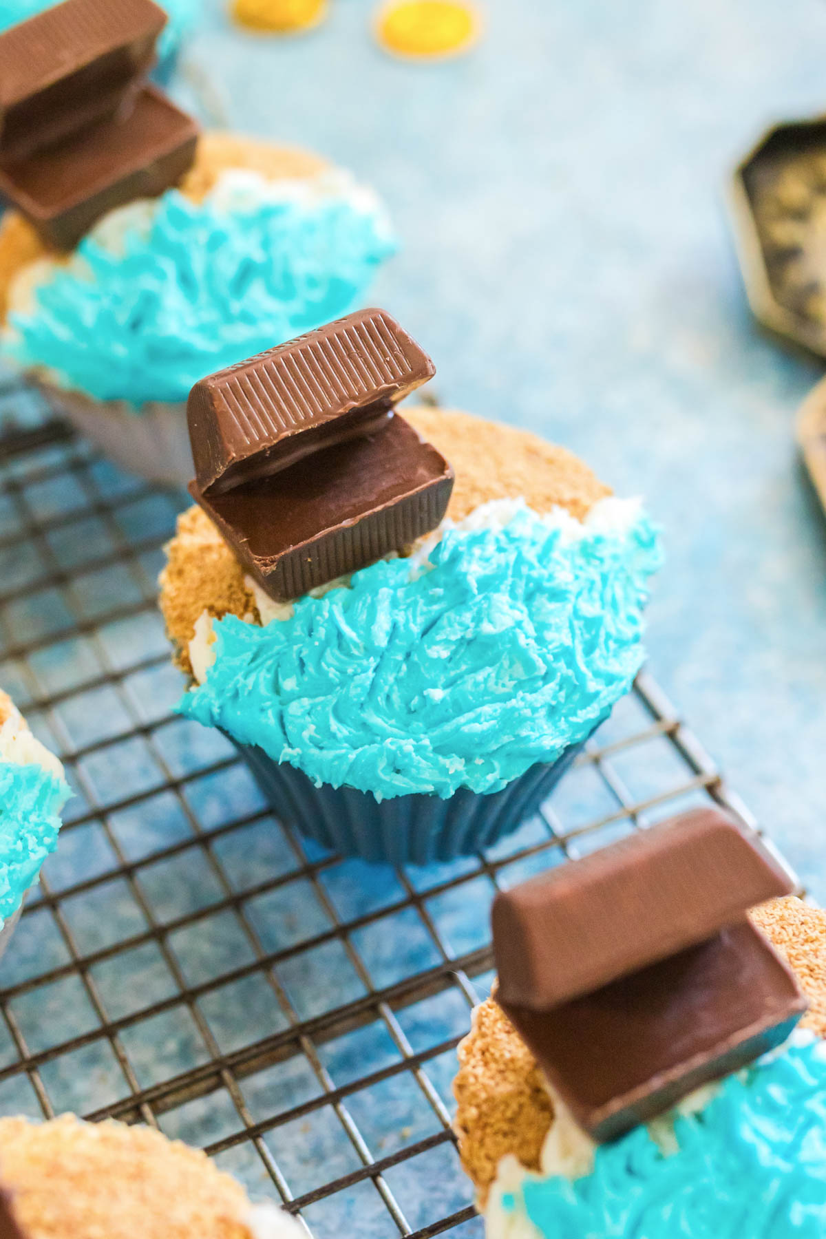 Cupcake decorated with blue frosting, crushed crumbs, and a chocolate piece shaped like a treasure chest, placed on a cooling rack.