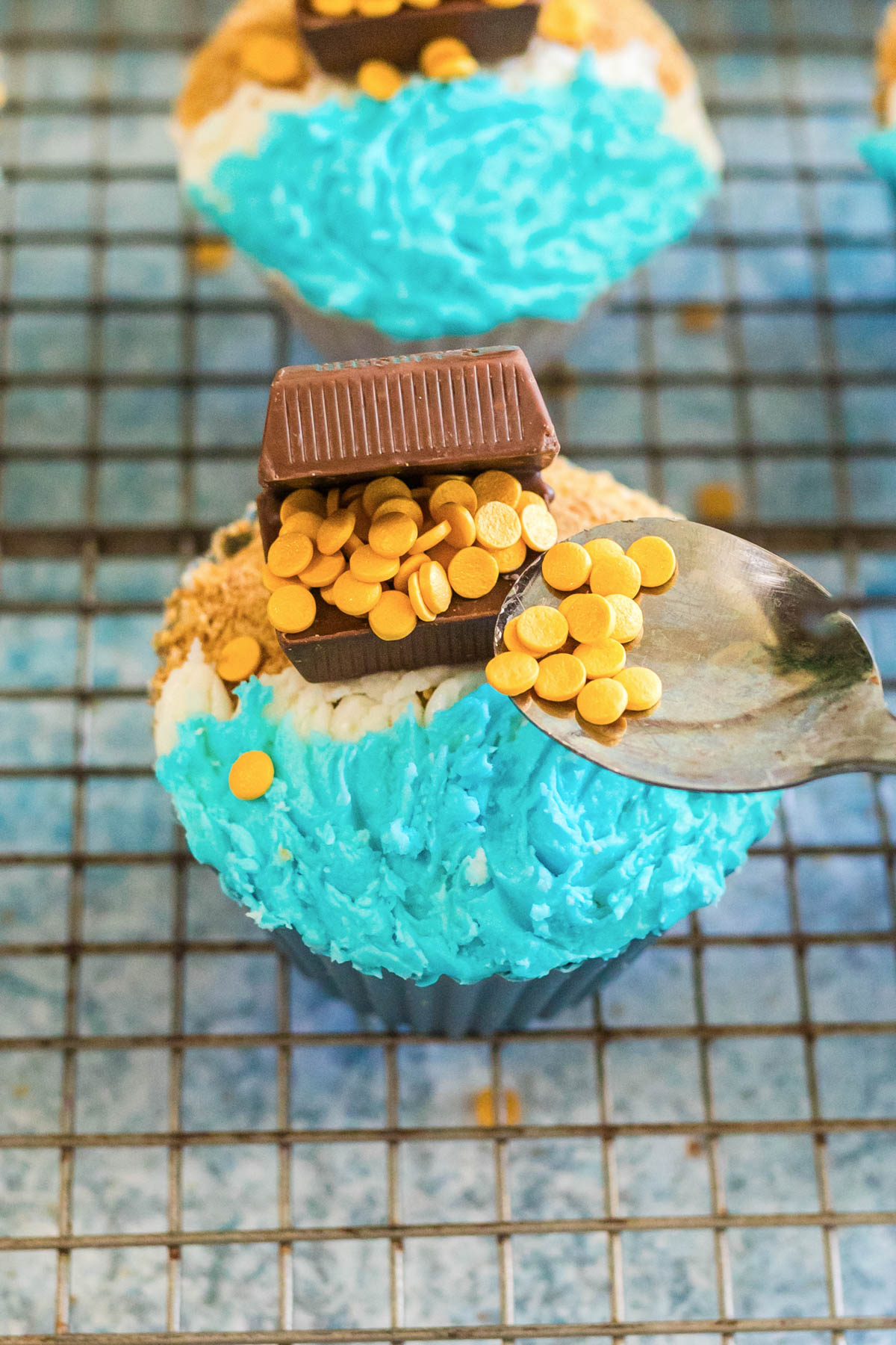 A cupcake decorated with blue and white frosting, topped with a chocolate treasure chest and gold coin sprinkles, sits on a wire cooling rack. A spoon adds more sprinkles.