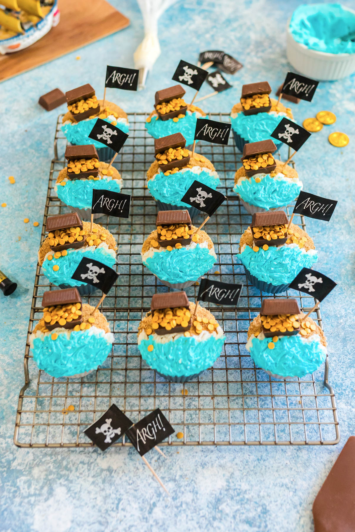 Cupcakes decorated with blue frosting, gold candy coins, chocolate pieces, and small pirate-themed flags are arranged on a wire cooling rack.