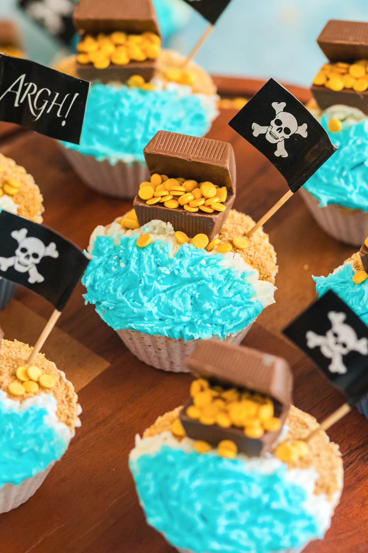 Cupcakes decorated with blue frosting to resemble water, topped with chocolate treasure chests filled with gold candy, and black pirate flags featuring a skull and crossbones.