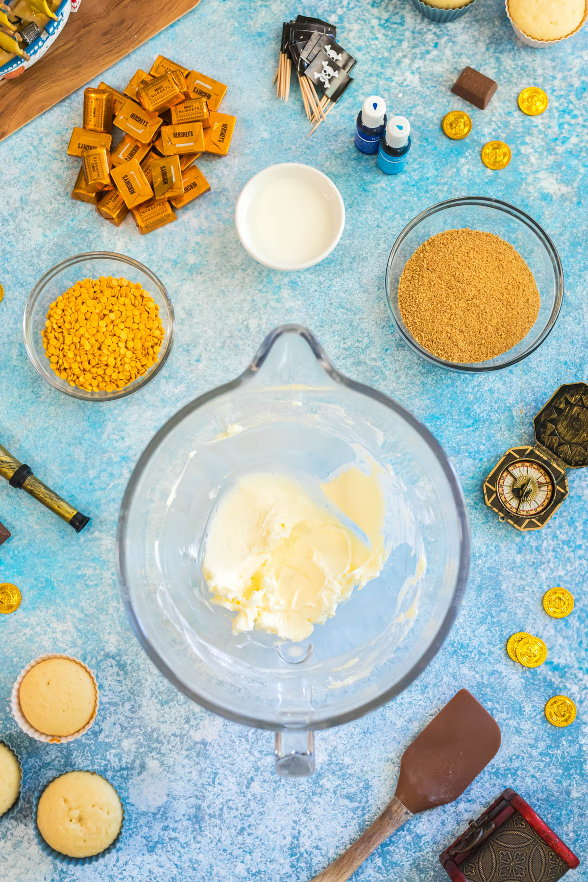 A blender with butter inside is surrounded by bowls of graham crumbs, yellow candy melts, and supplies on a blue countertop.