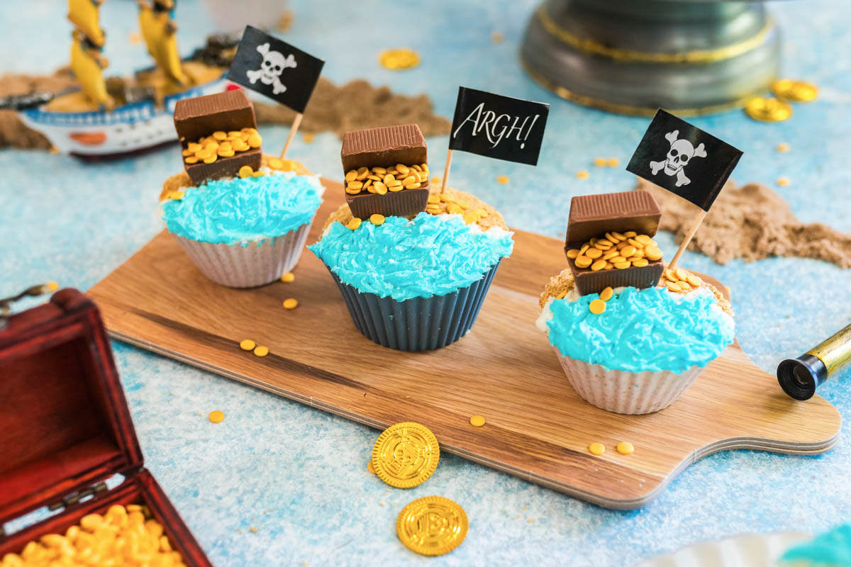 Three cupcakes decorated with blue frosting, chocolate pieces, candy gold coins, and pirate-themed flags on a wooden tray; gold coins and pirate props surround them.