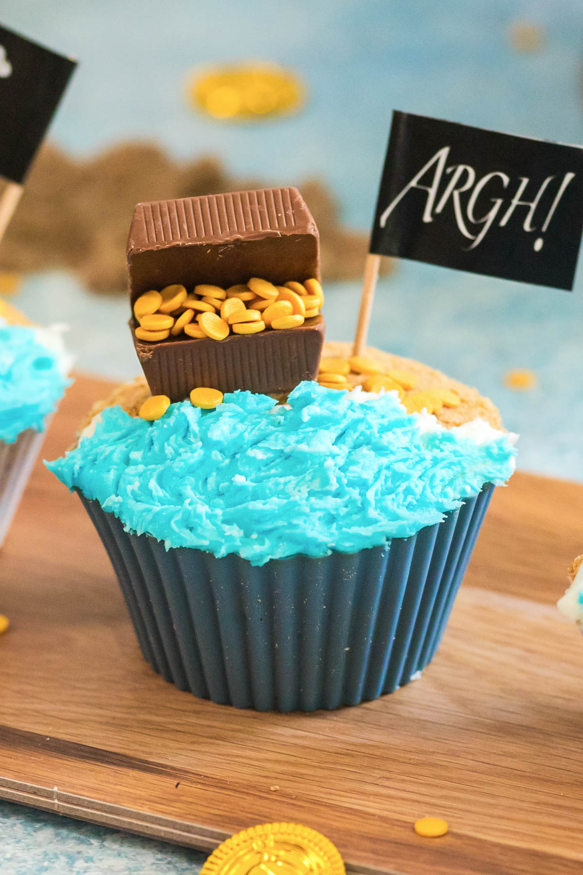 A cupcake decorated with blue icing to resemble water, topped with a chocolate treasure chest filled with gold candy and a small flag reading "ARGH!".