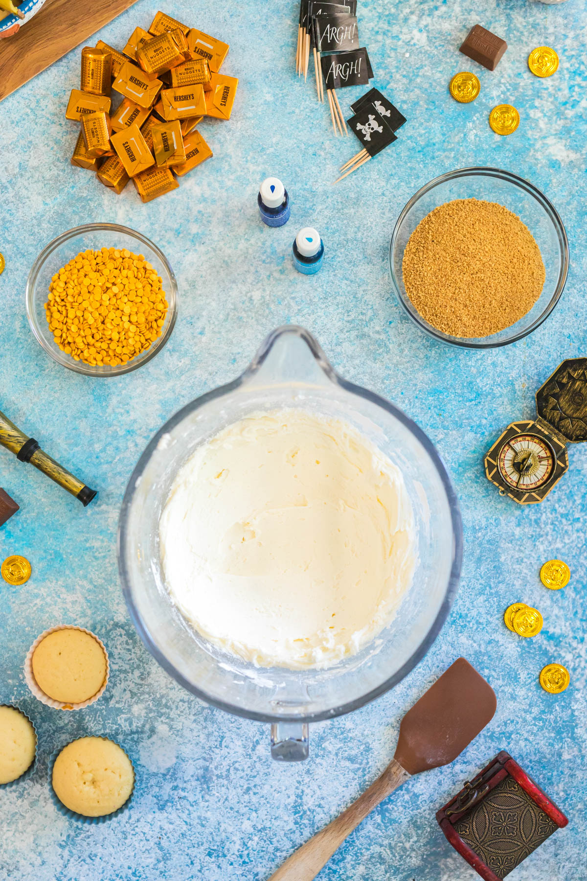 A mixing bowl with butter and powdered sugar filling in the center, surrounded by bowls of yellow sprinkles, graham crumbs, chocolates, cupcakes, pirate-themed props, and blue food coloring on a blue surface.