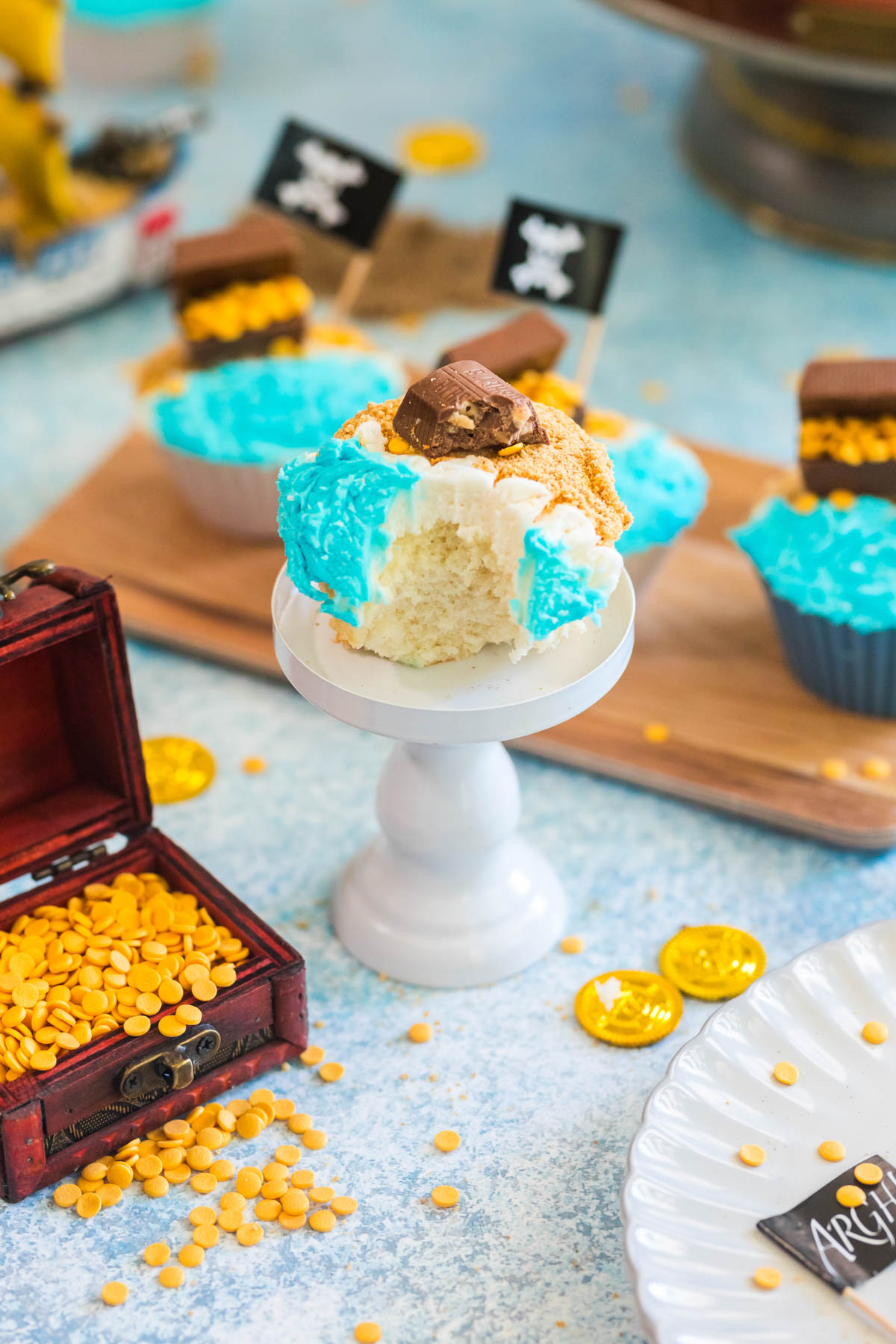A partially eaten cupcake with blue and white frosting and a chocolate piece on top sits on a small white stand. Pirate-themed decorations and gold coins are in the background.