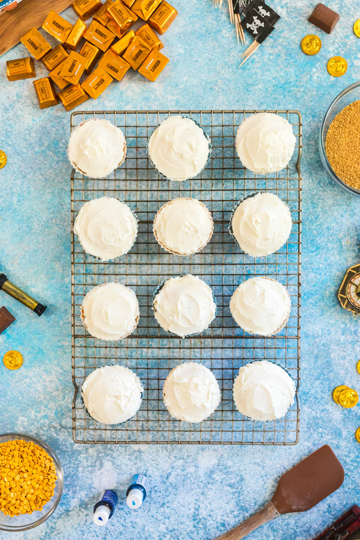 Twelve cupcakes with white frosting arranged on a wire cooling rack, surrounded by baking ingredients and gold-wrapped candies on a blue surface.