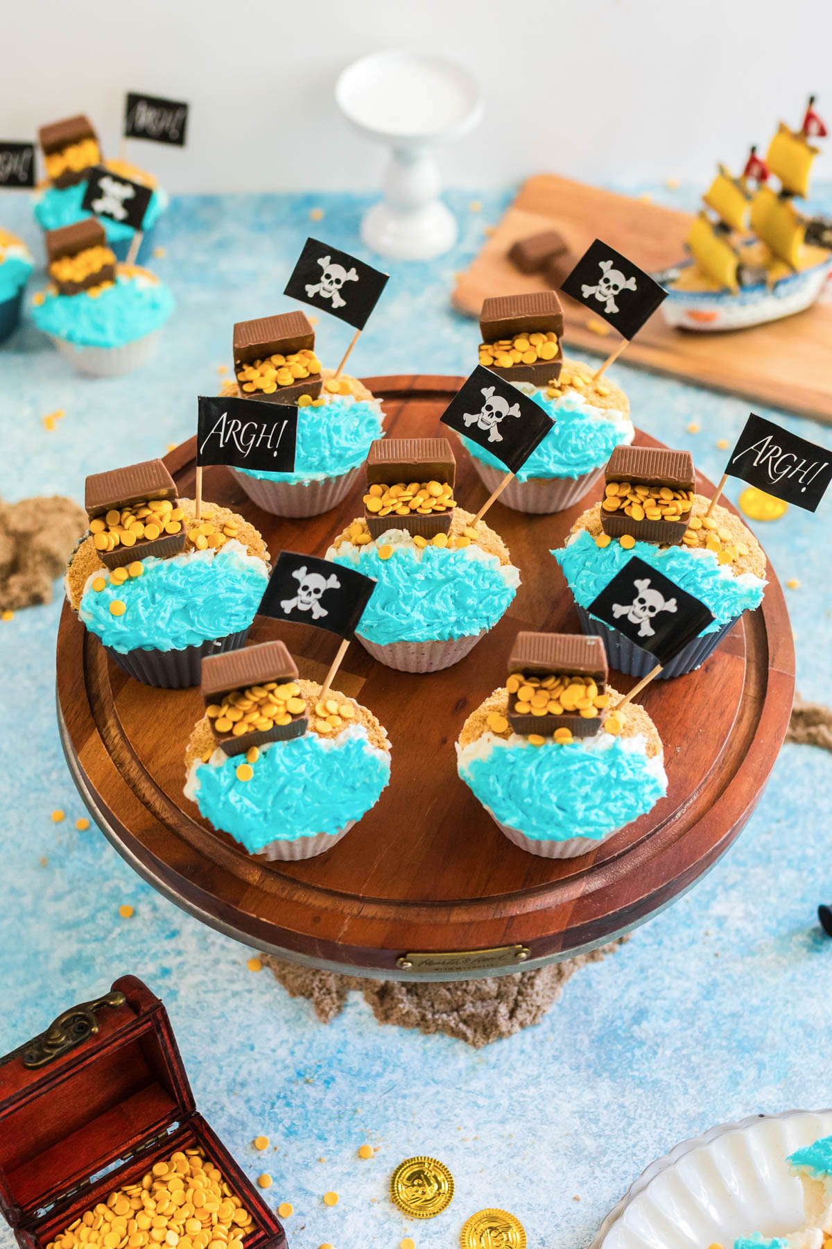 Pirate-themed cupcakes with blue frosting, chocolate pieces, gold sprinkles, and small skull-and-crossbones flags on a wooden stand; a toy ship and treasure chest are in the background.