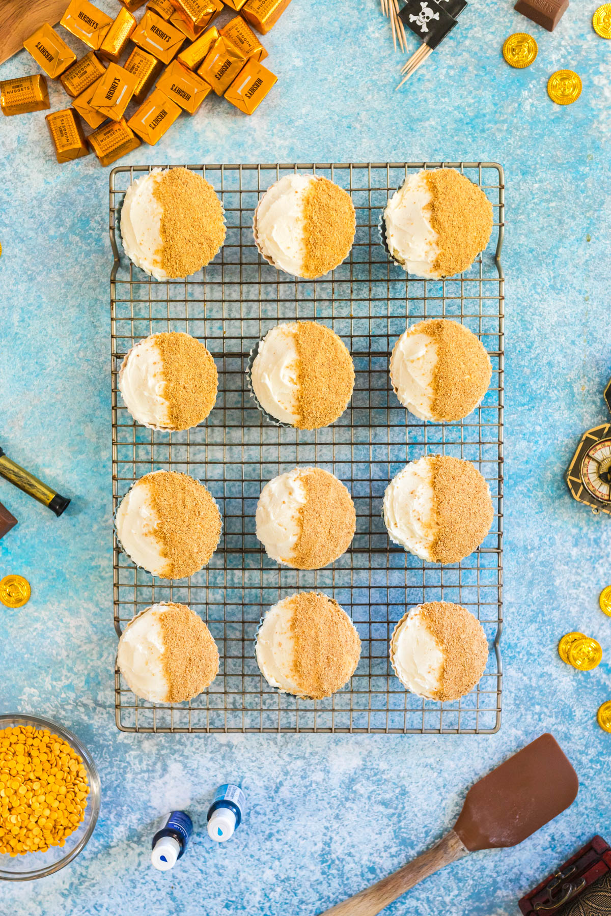 Twelve cupcakes with white frosting and half covered in graham cracker crumbs are arranged on a wire rack, surrounded by gold coins, mini chocolate bars, and baking tools on a blue surface.