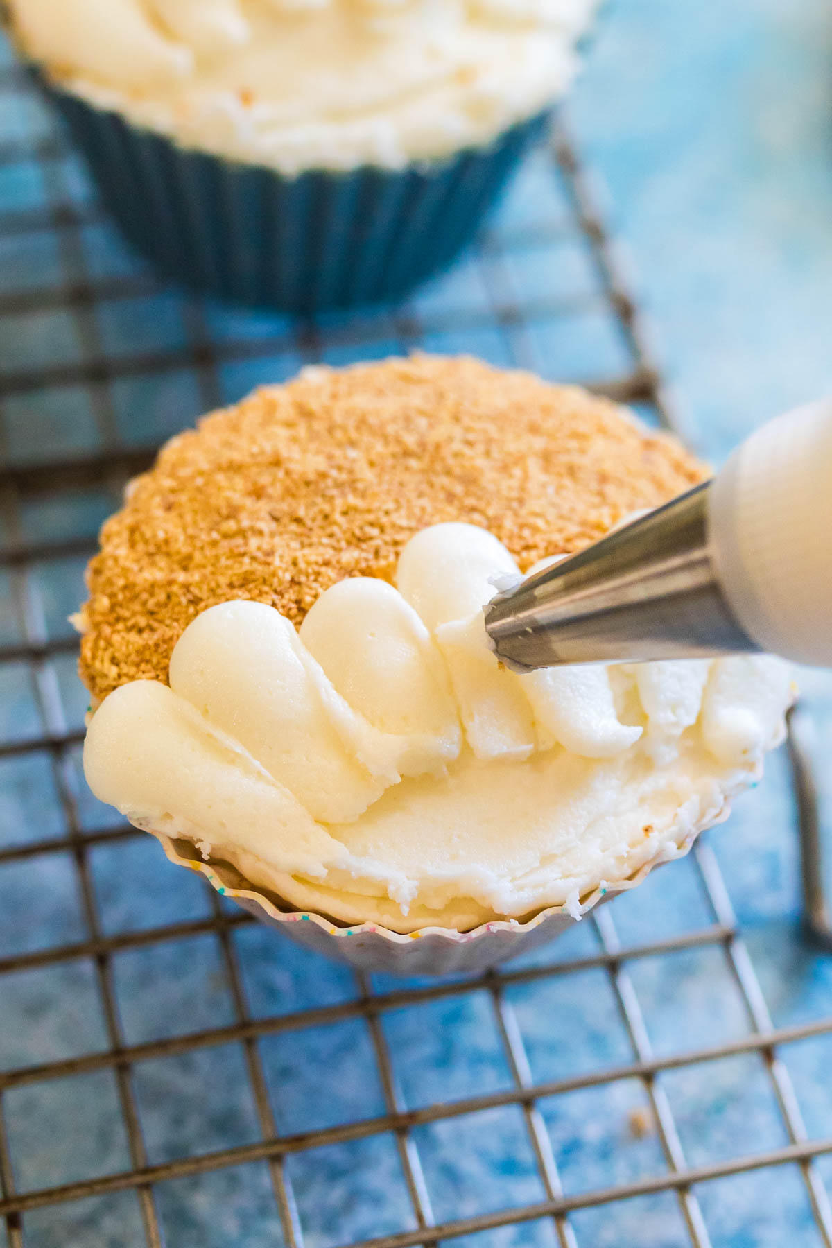 A cupcake is being decorated with white frosting using a piping bag. The cupcake sits on a metal cooling rack, and has a crumb topping on one half.