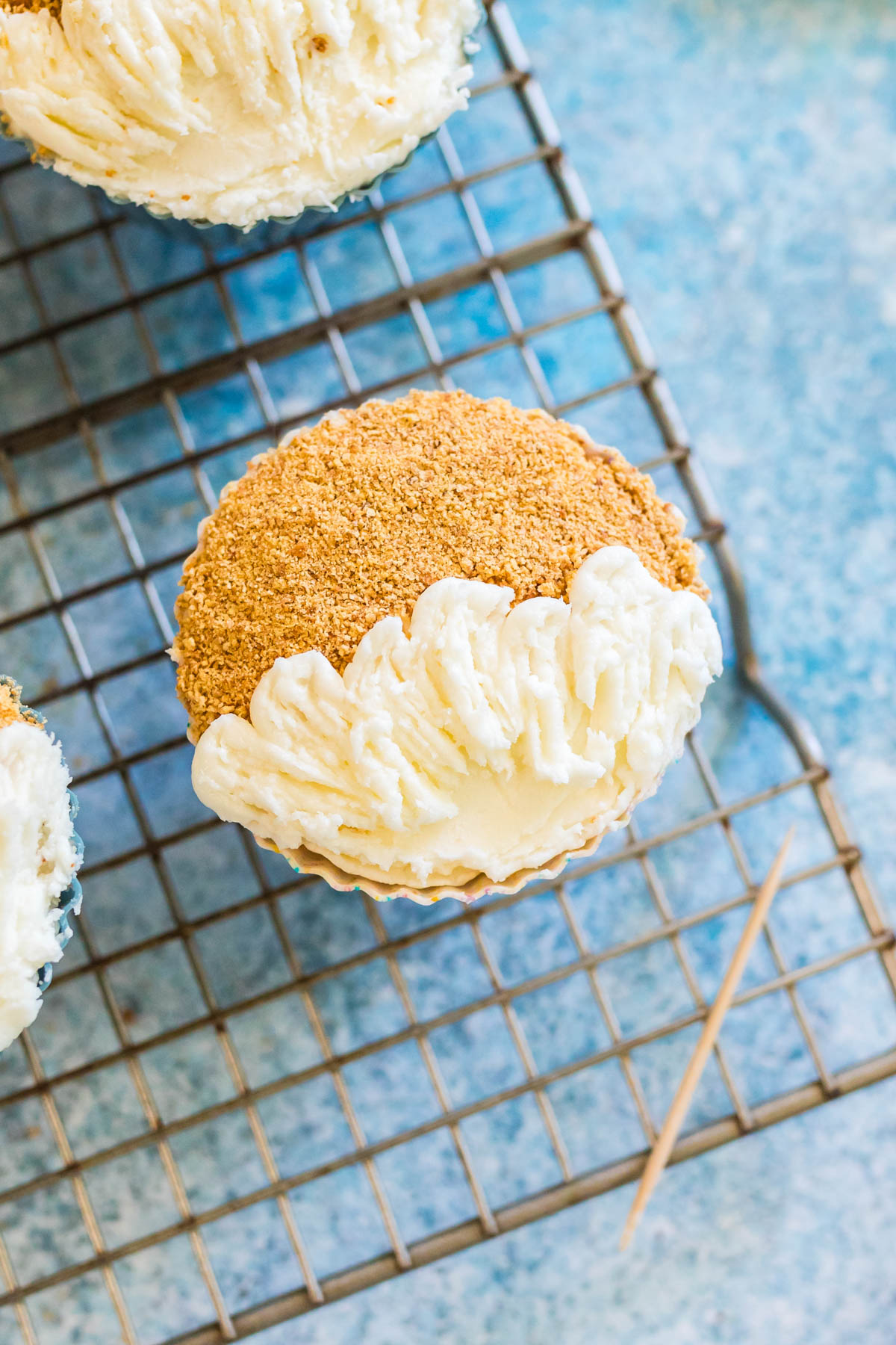 A cupcake with white frosting and crushed graham cracker topping sits on a cooling rack with a toothpick nearby.