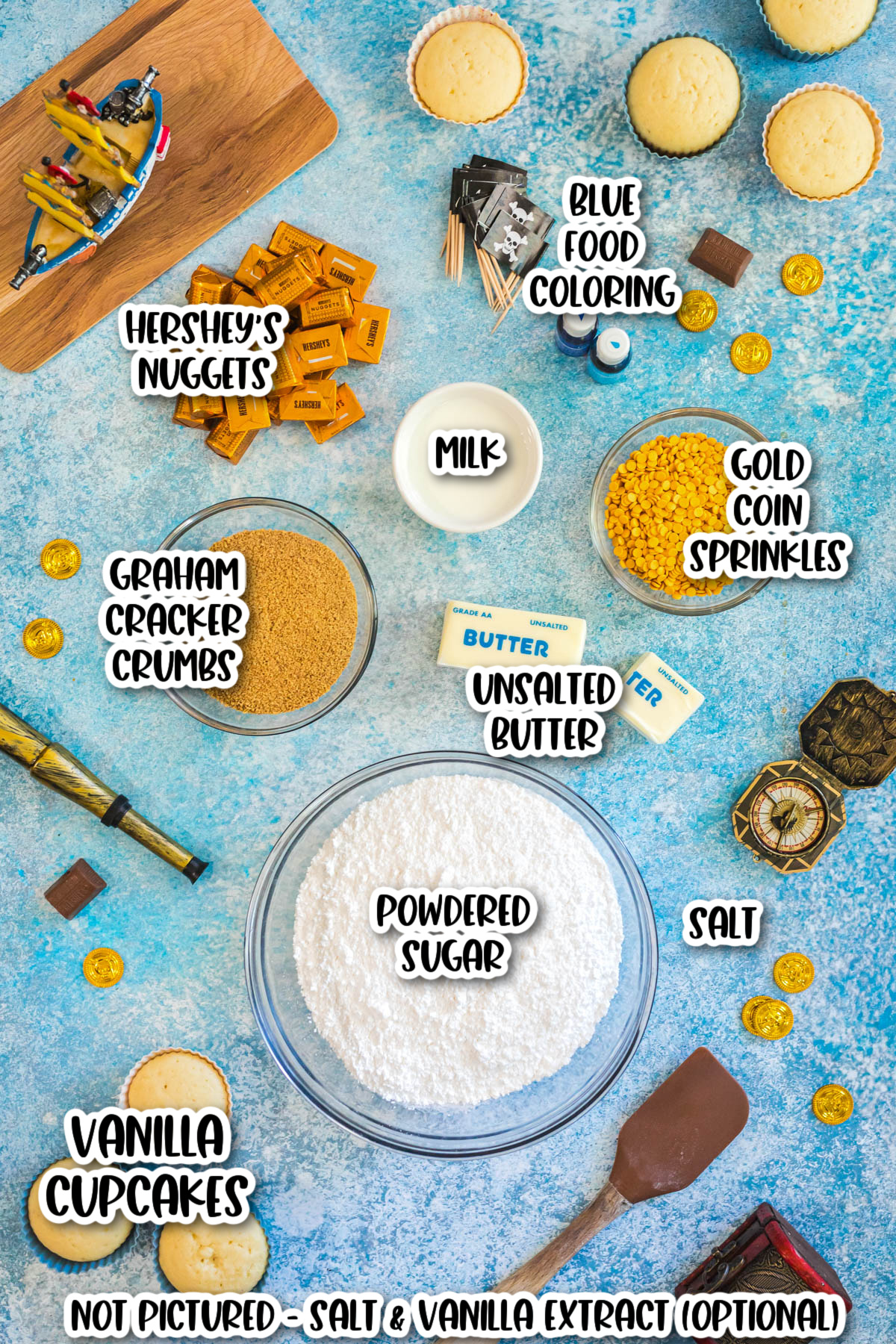 Flat lay of baking ingredients for cupcakes, including vanilla cupcakes, powdered sugar, butter, milk, graham cracker crumbs, Hershey&rsquo;s Nuggets, gold coin sprinkles, salt, and blue food coloring.