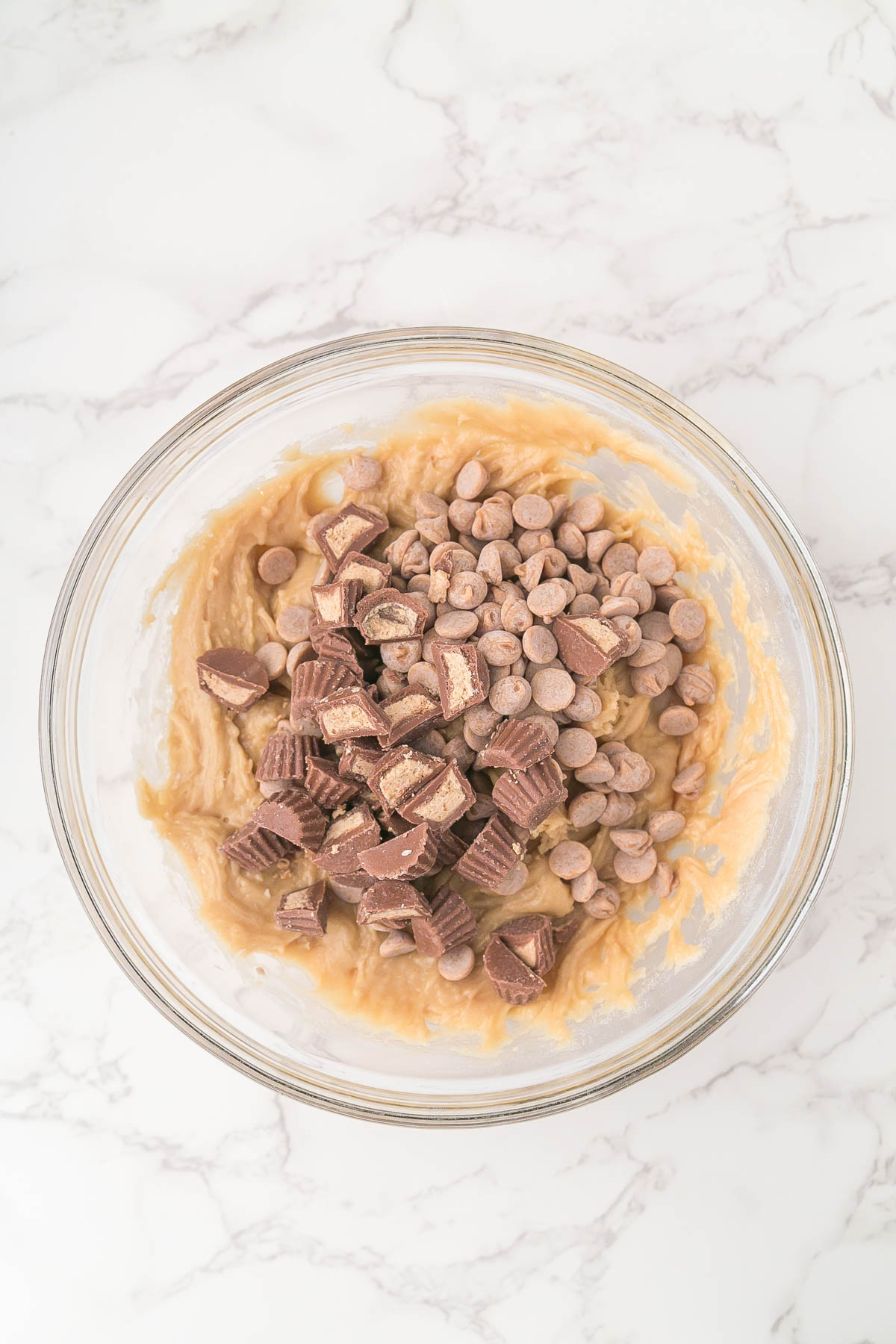 A glass bowl filled with cookie dough, peanut butter chips, and chopped peanut butter cups on a white marble surface.