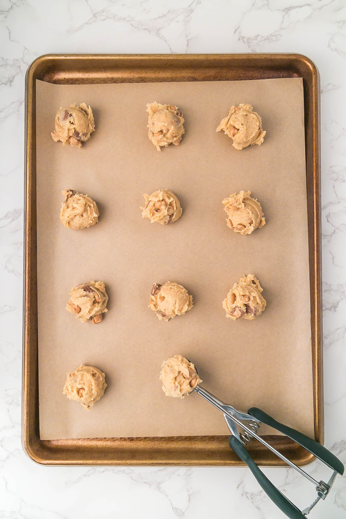 A baking sheet lined with parchment paper holds twelve evenly spaced scoops of cookie dough. An ice cream scoop with dough is placed in the lower right corner of the tray.