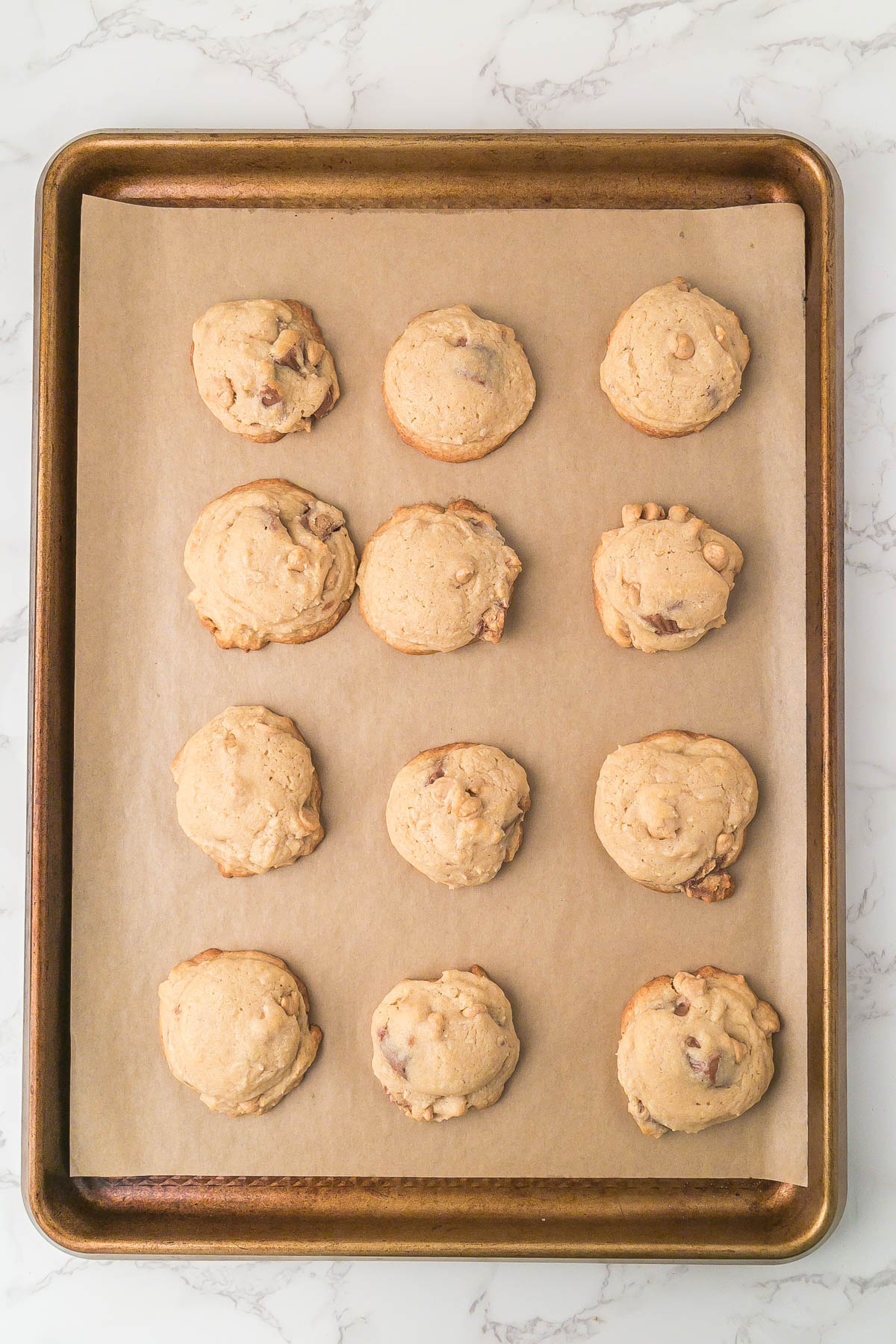 A baking sheet lined with parchment paper holds twelve round, baked cookies spaced evenly in rows of three on a white marble surface.