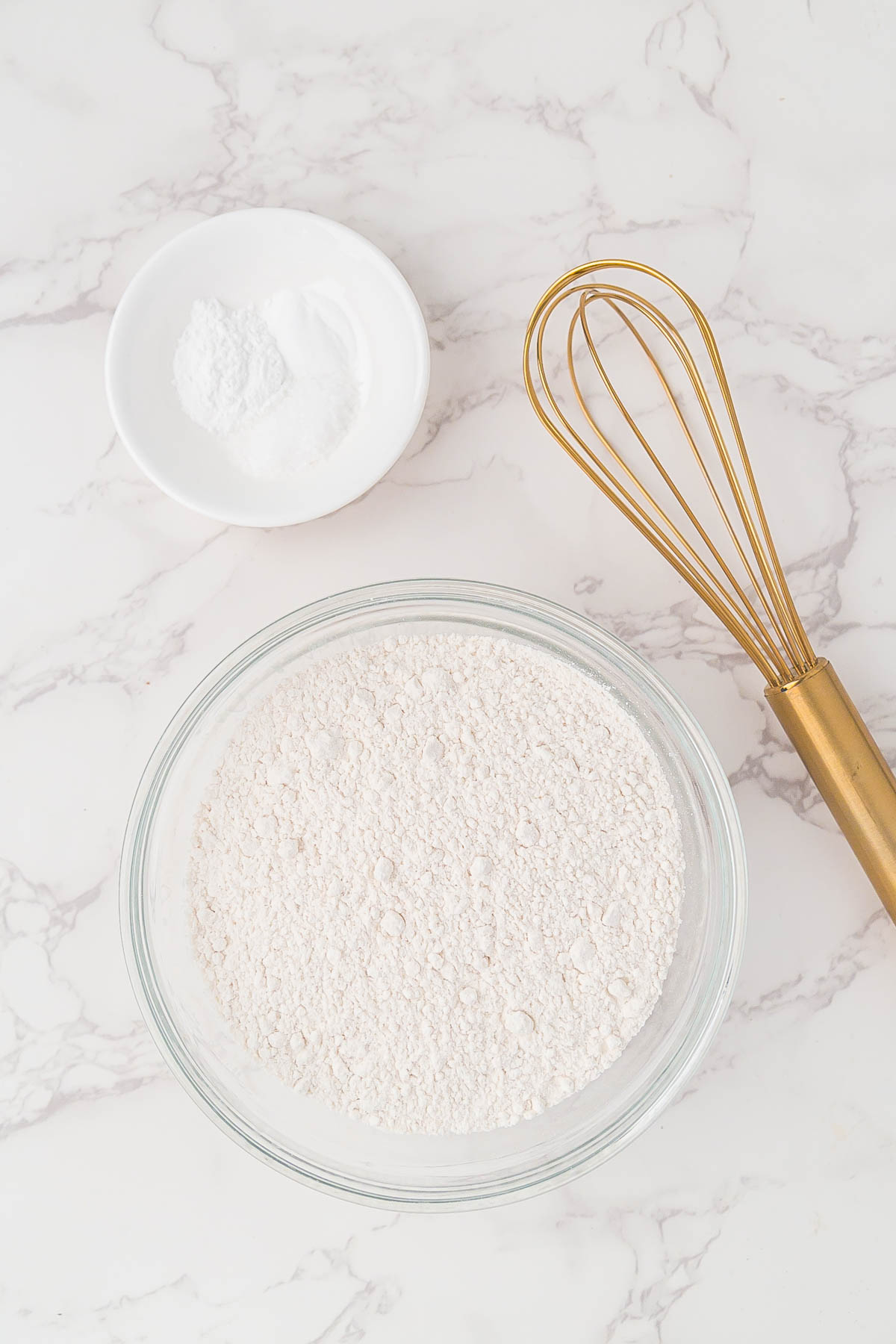 A glass bowl filled with flour, a small white dish holding baking powder, and a gold metal whisk arranged on a marble surface.
