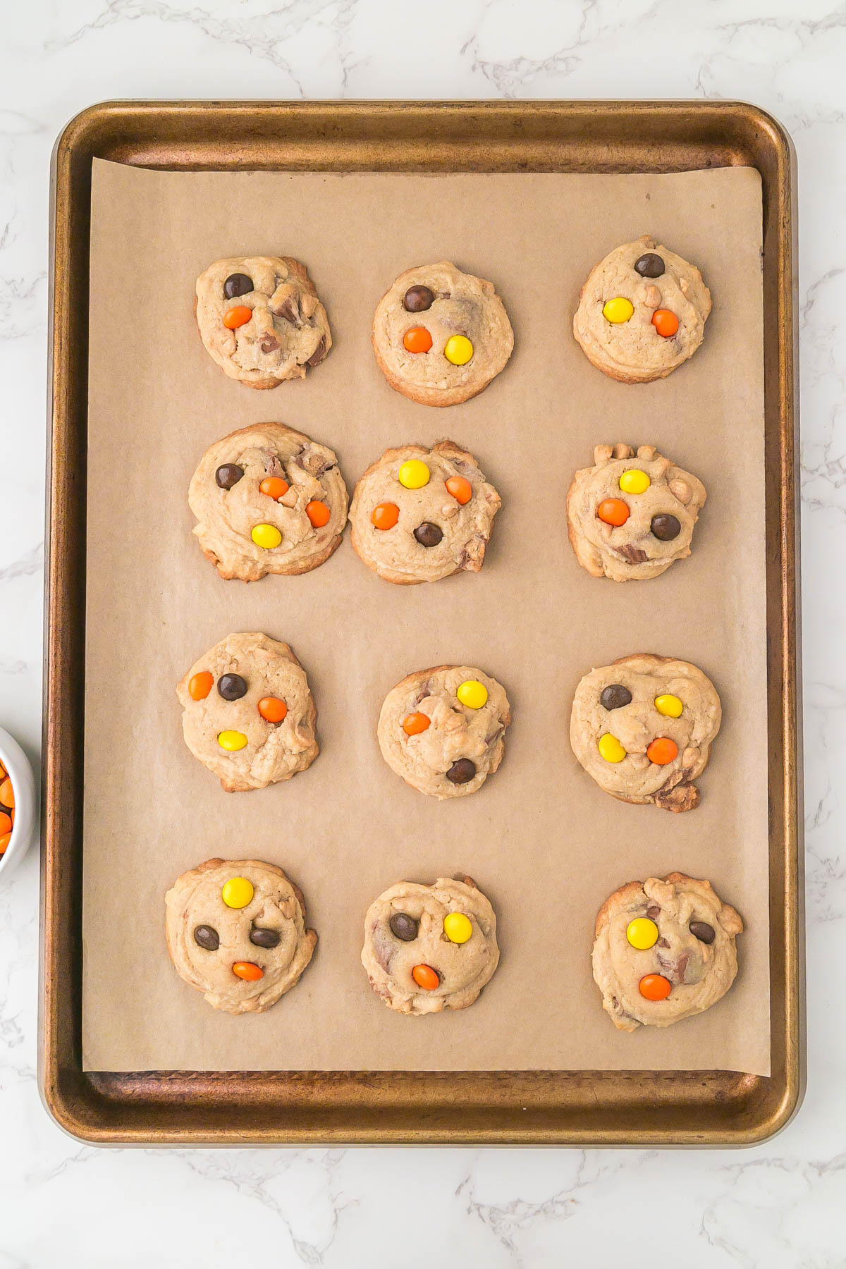 A baking sheet lined with parchment paper holds twelve unbaked cookie dough balls topped with yellow, orange, and brown candy-coated chocolates.