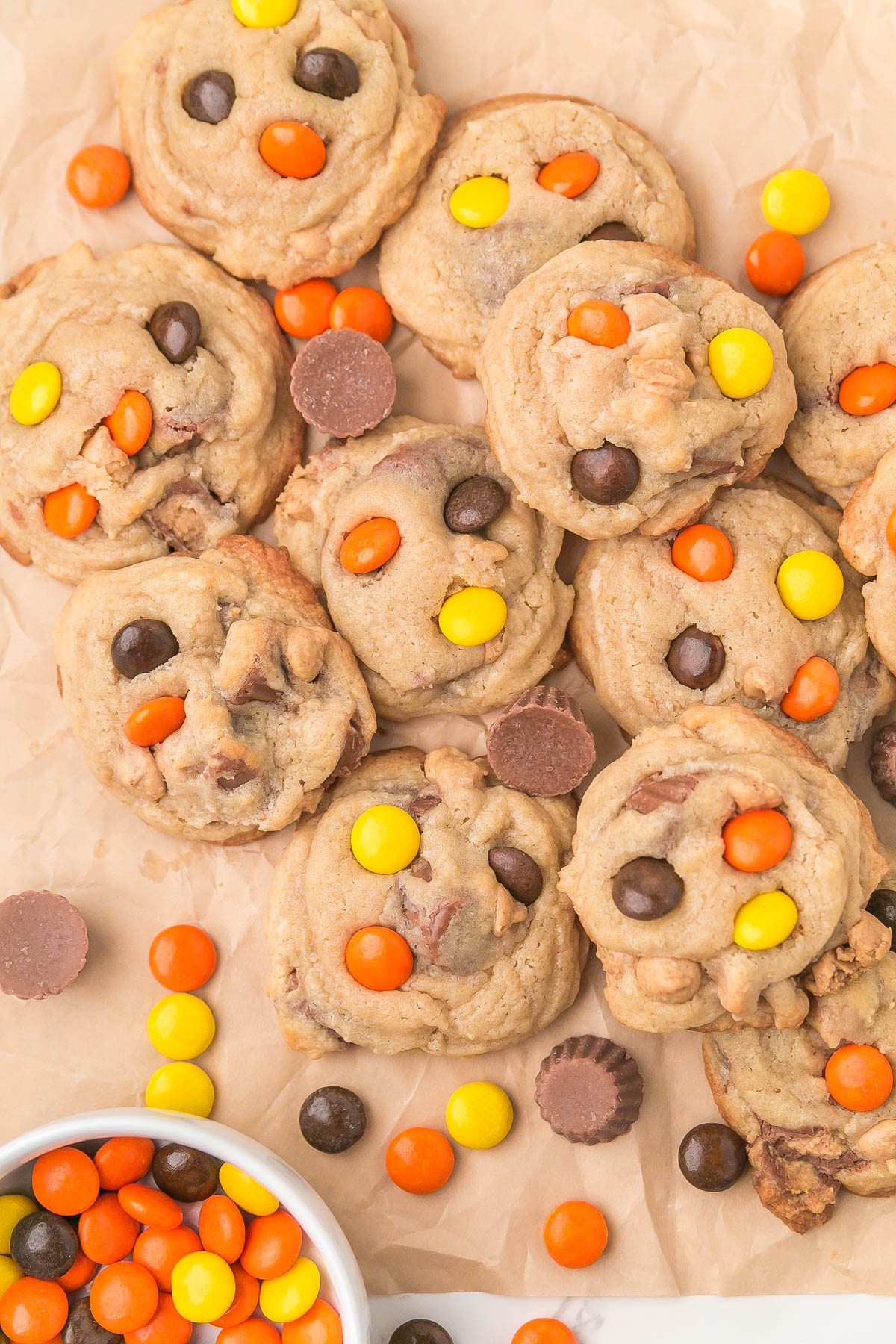 A batch of peanut butter cookies topped with orange, yellow, and brown candy-coated chocolates and mini peanut butter cups on parchment paper.