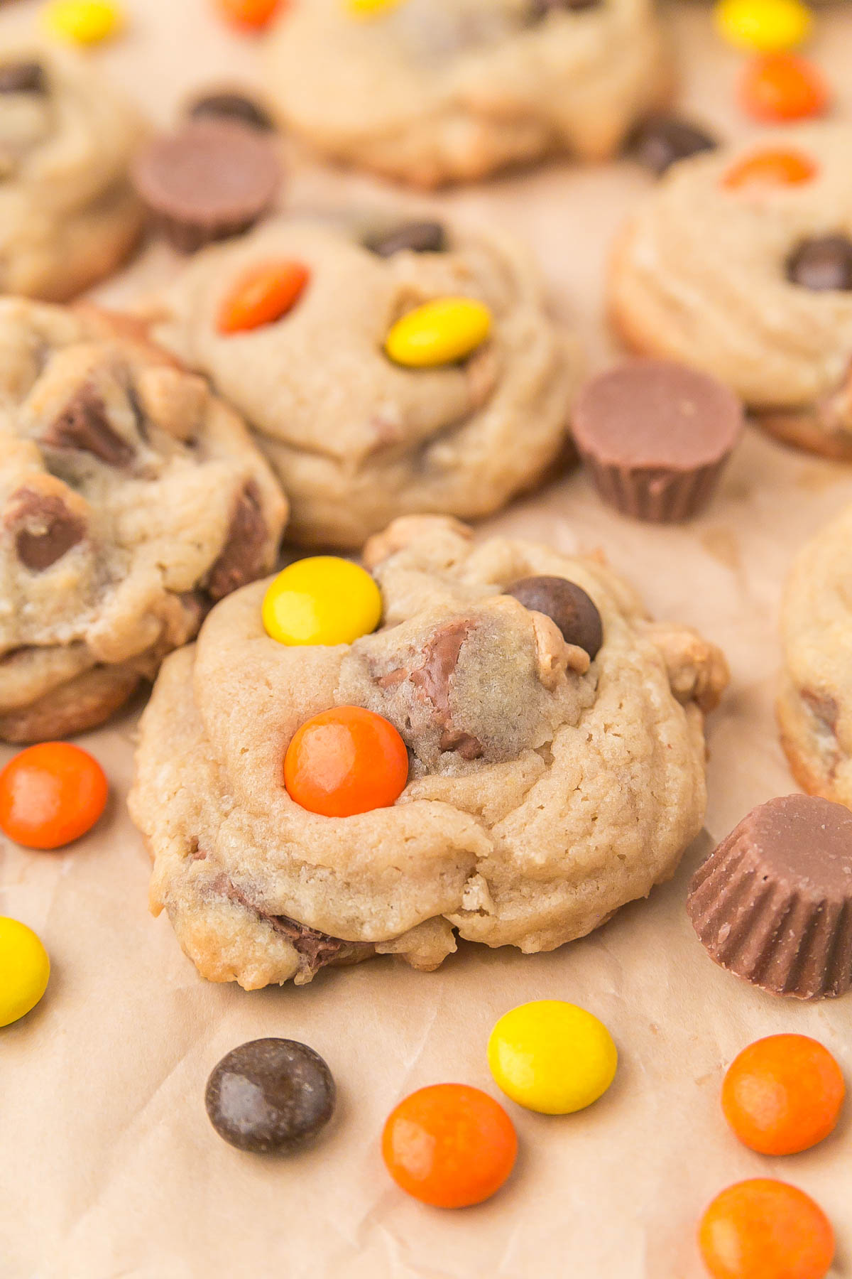 Peanut butter cookies with chocolate chips and colorful candy-coated chocolates, surrounded by additional candies and mini peanut butter cups on parchment paper.
