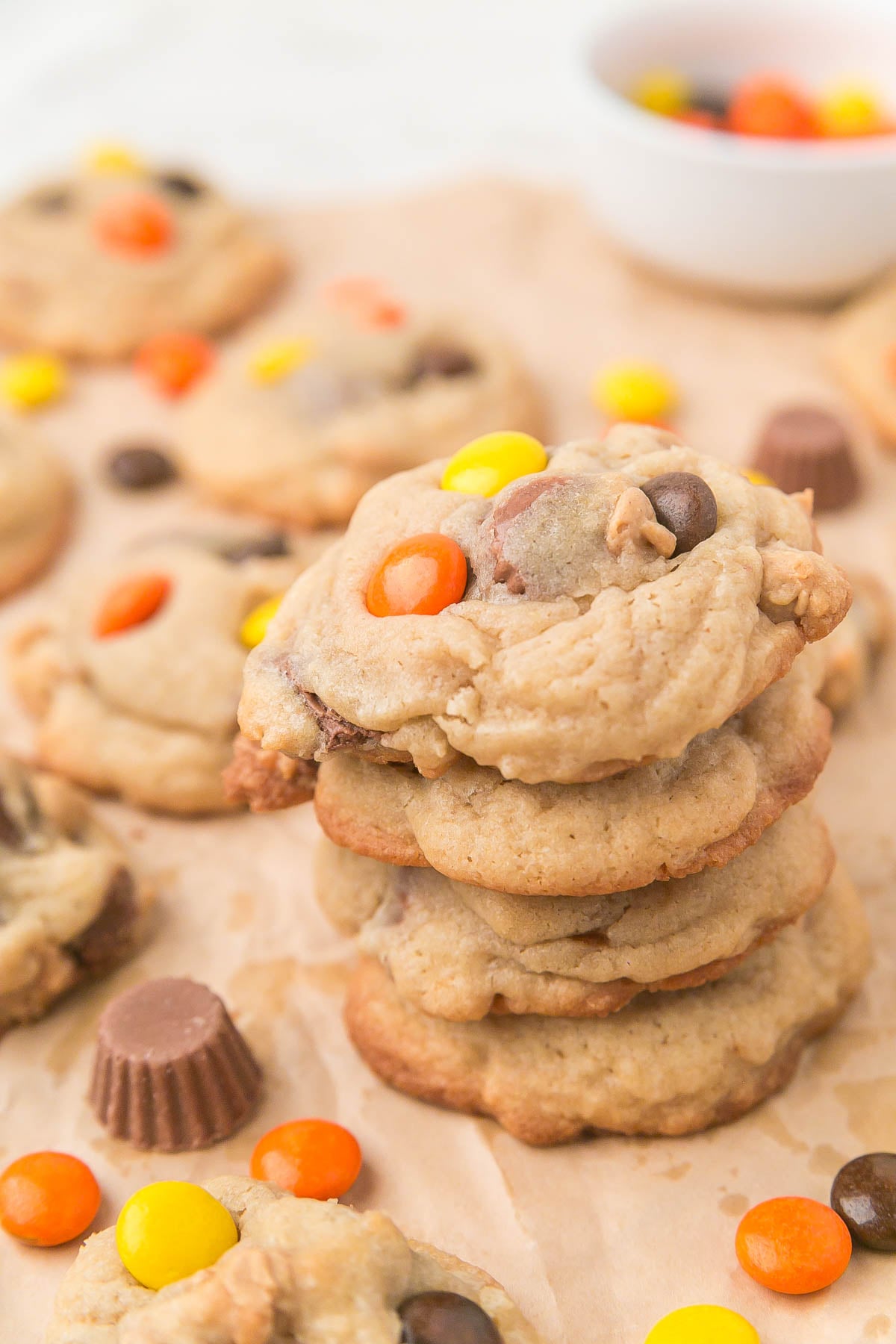 A stack of cookies with chocolate chips and colorful candy-coated chocolates, surrounded by more cookies and candies on a parchment-lined surface.