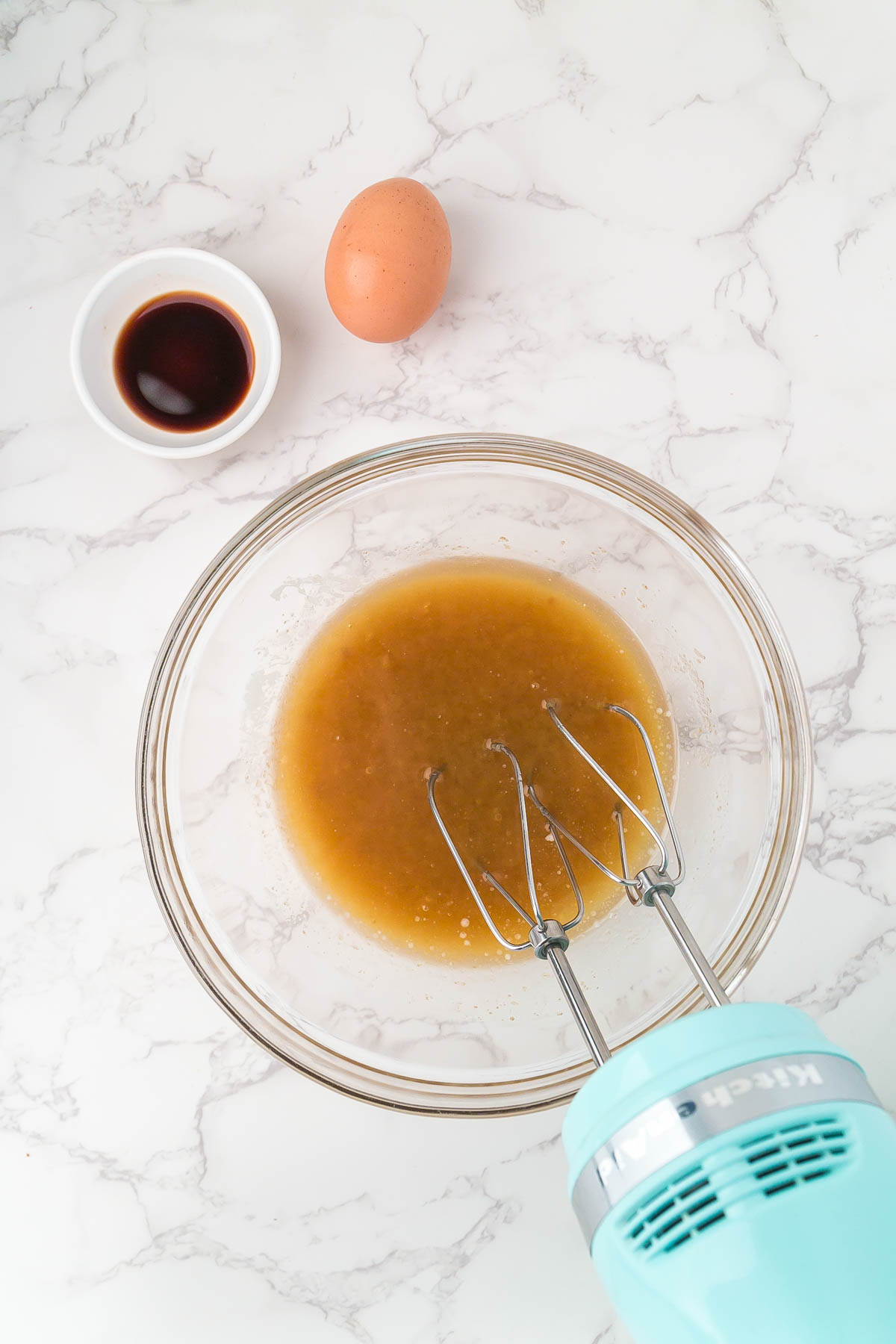 A glass bowl with brown liquid mixture being mixed by a hand mixer, next to an egg and a small bowl of vanilla extract on a marble surface.