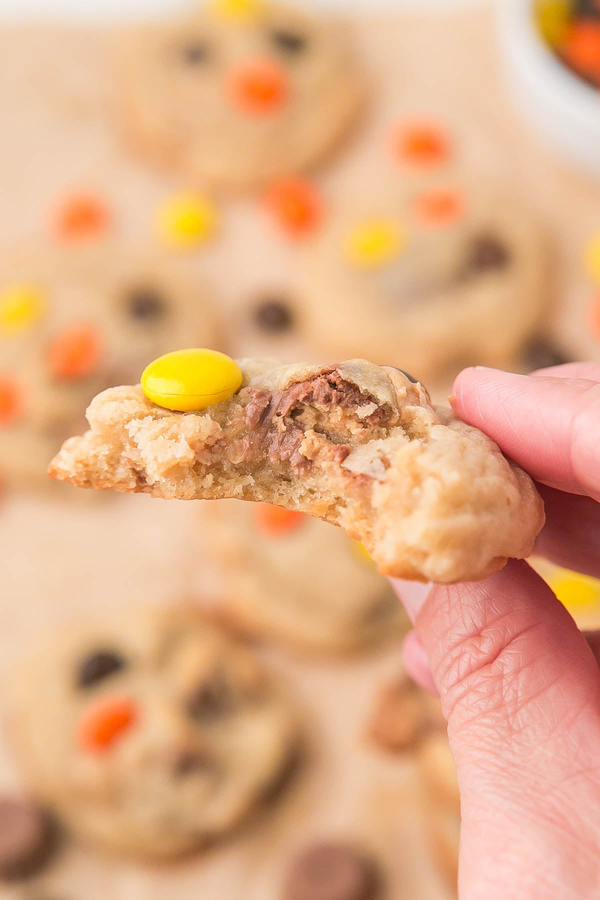 A hand holding a partially eaten cookie topped with a yellow candy-coated chocolate, with more cookies in the blurred background.