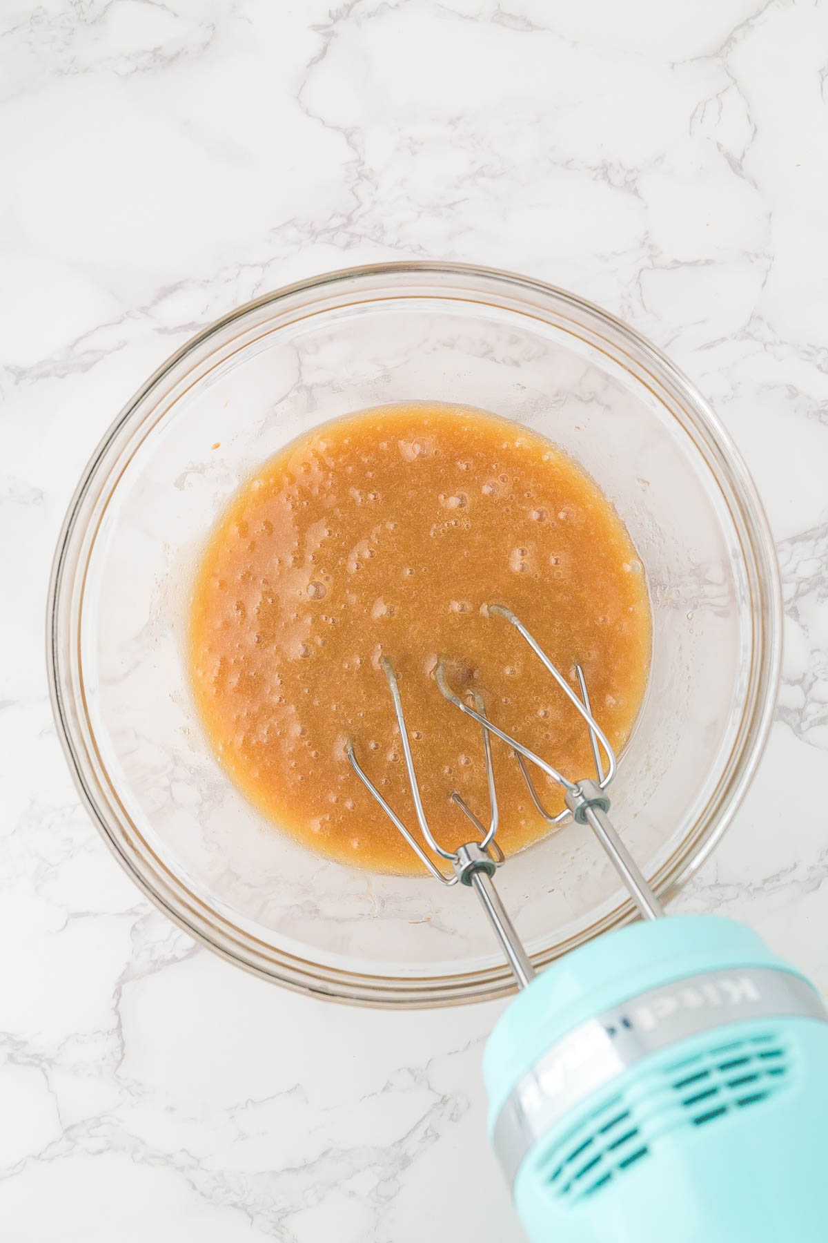 A glass bowl containing a light brown mixture being blended with an electric hand mixer on a white marble surface.