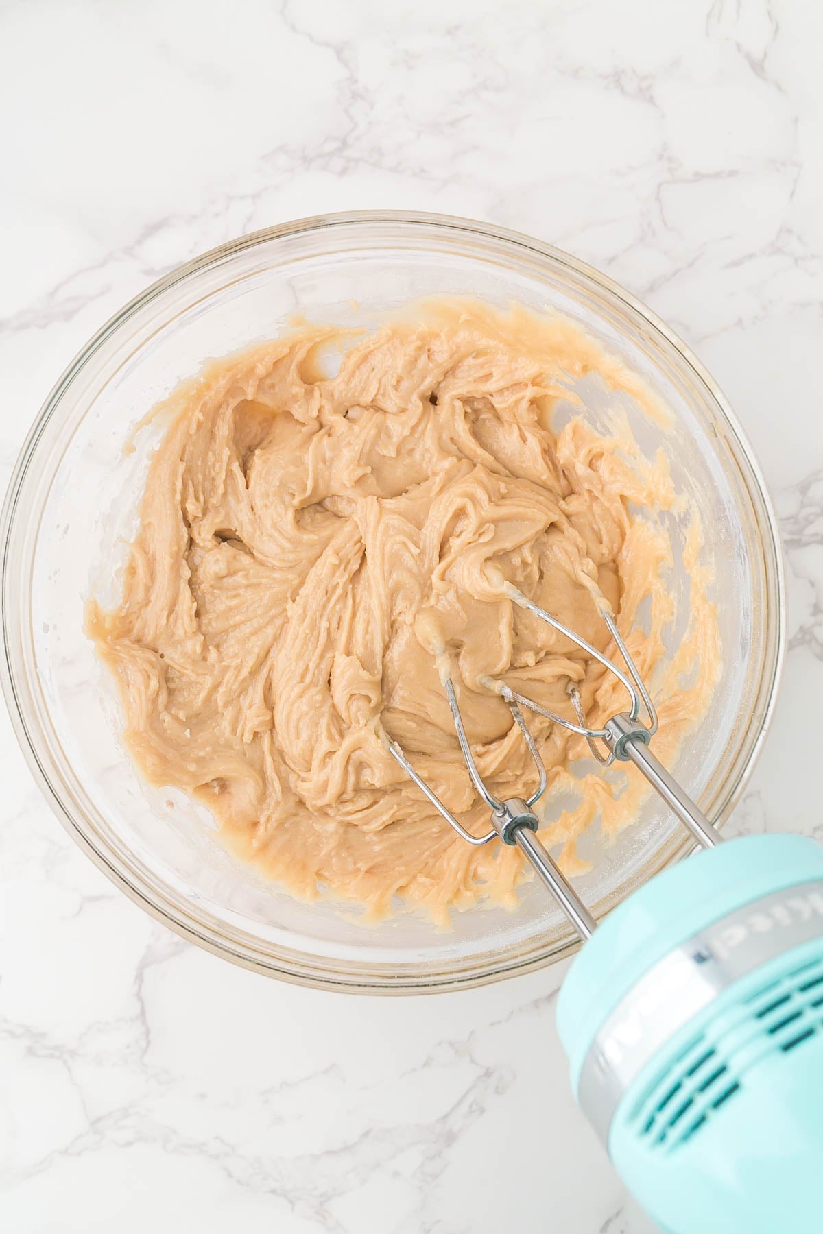 A glass bowl containing light brown batter being mixed with an electric hand mixer on a marble countertop.