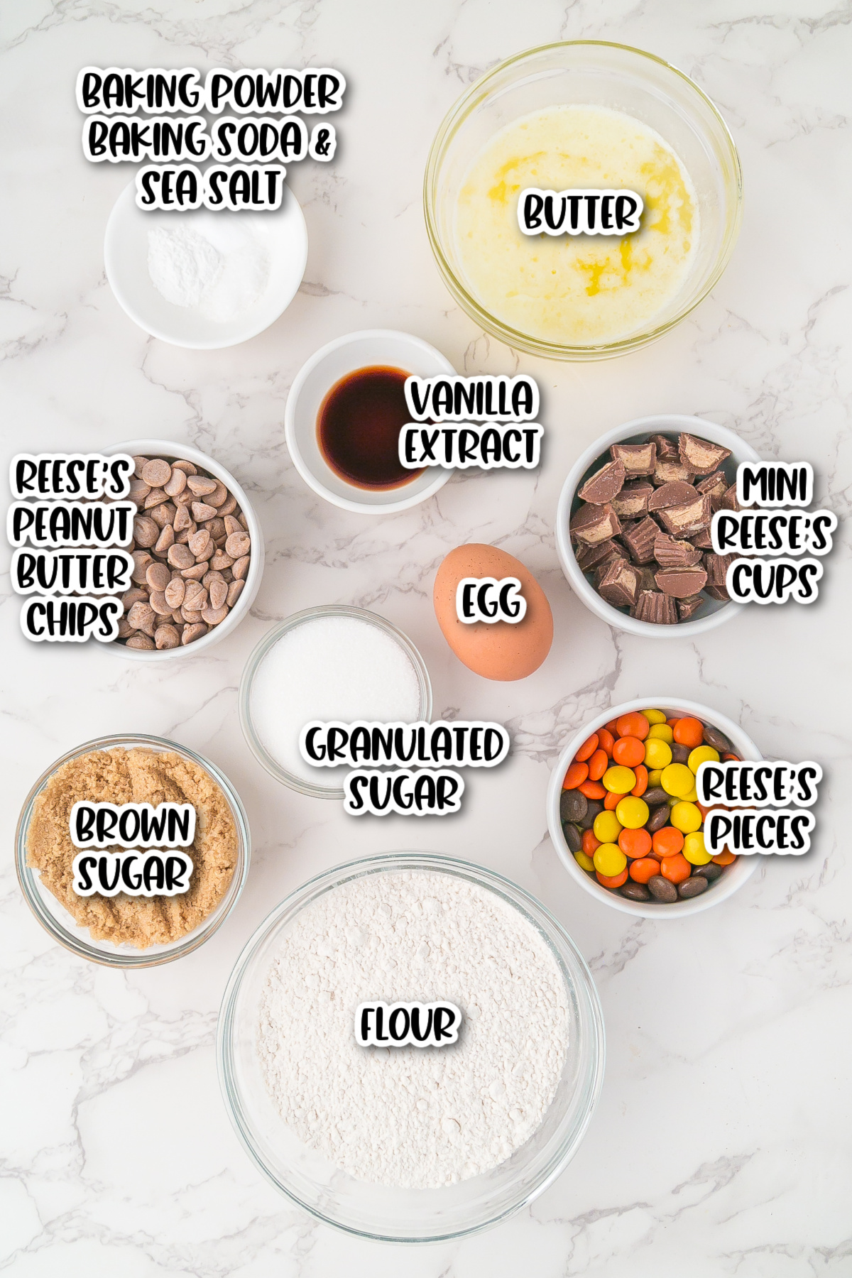 Baking ingredients arranged on a marble surface, including flour, sugars, butter, egg, vanilla, baking powder, salt, peanut butter chips, mini peanut butter cups, and Reese's Pieces.