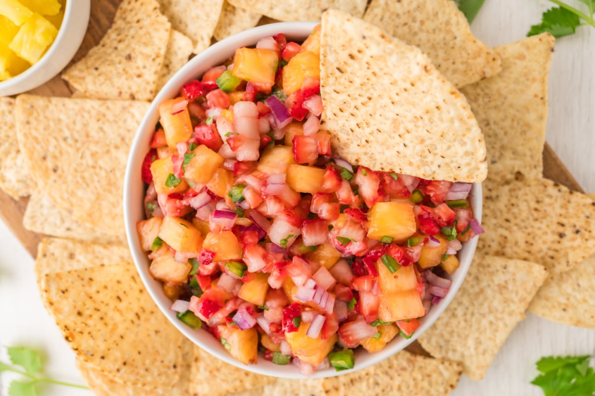 A bowl of colorful fruit salsa with diced pineapple, red onion, and herbs, surrounded by tortilla chips, with one chip dipped in the salsa.