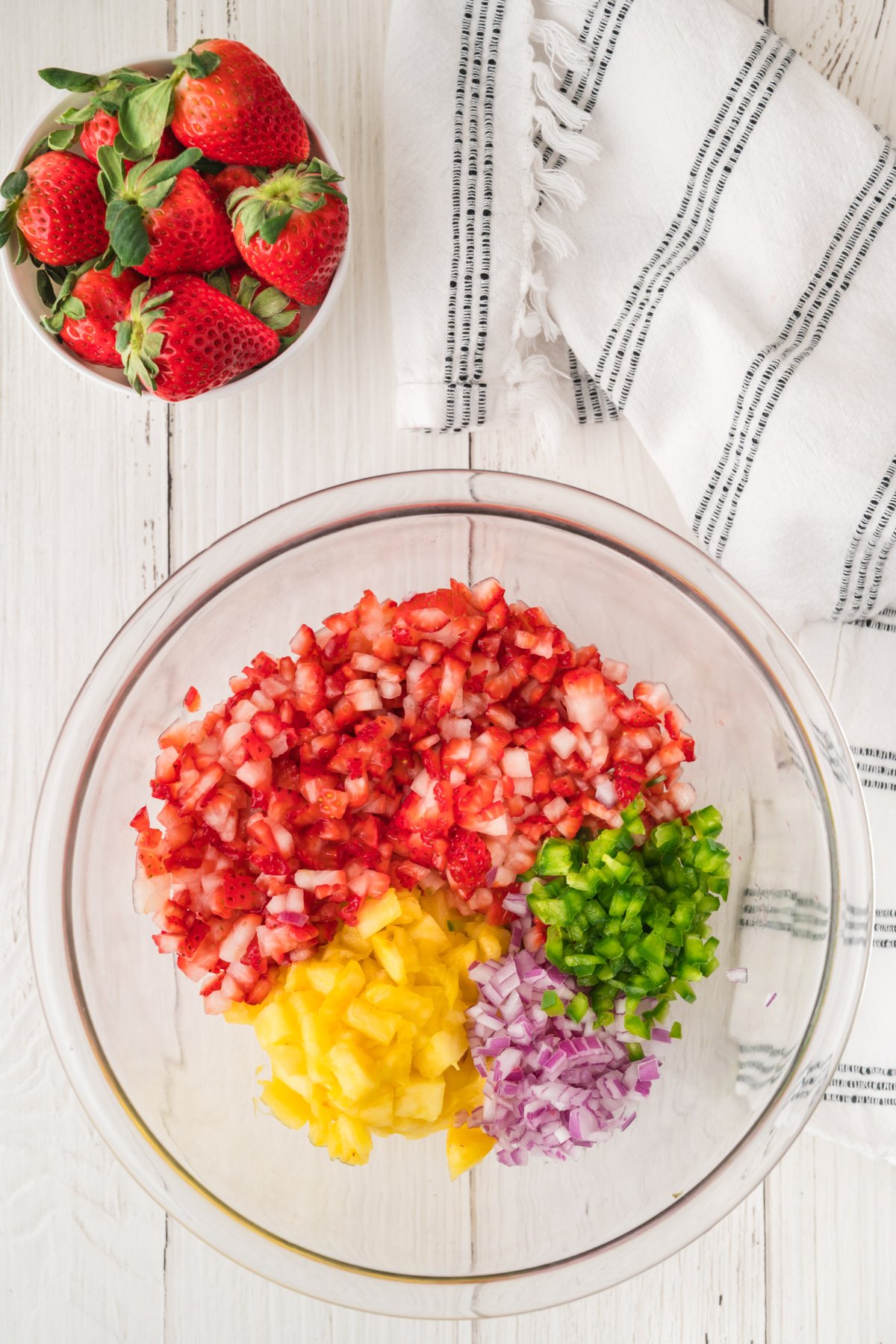 A glass bowl contains chopped strawberries, pineapple, red onion, and jalape&ntilde;o. A bowl of whole strawberries and a white striped towel are nearby on a white wooden surface.