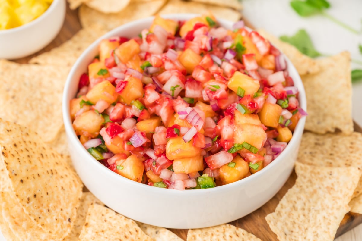 A white bowl filled with colorful salsa made of diced pineapple, red onion, cilantro, and peppers, surrounded by tortilla chips.