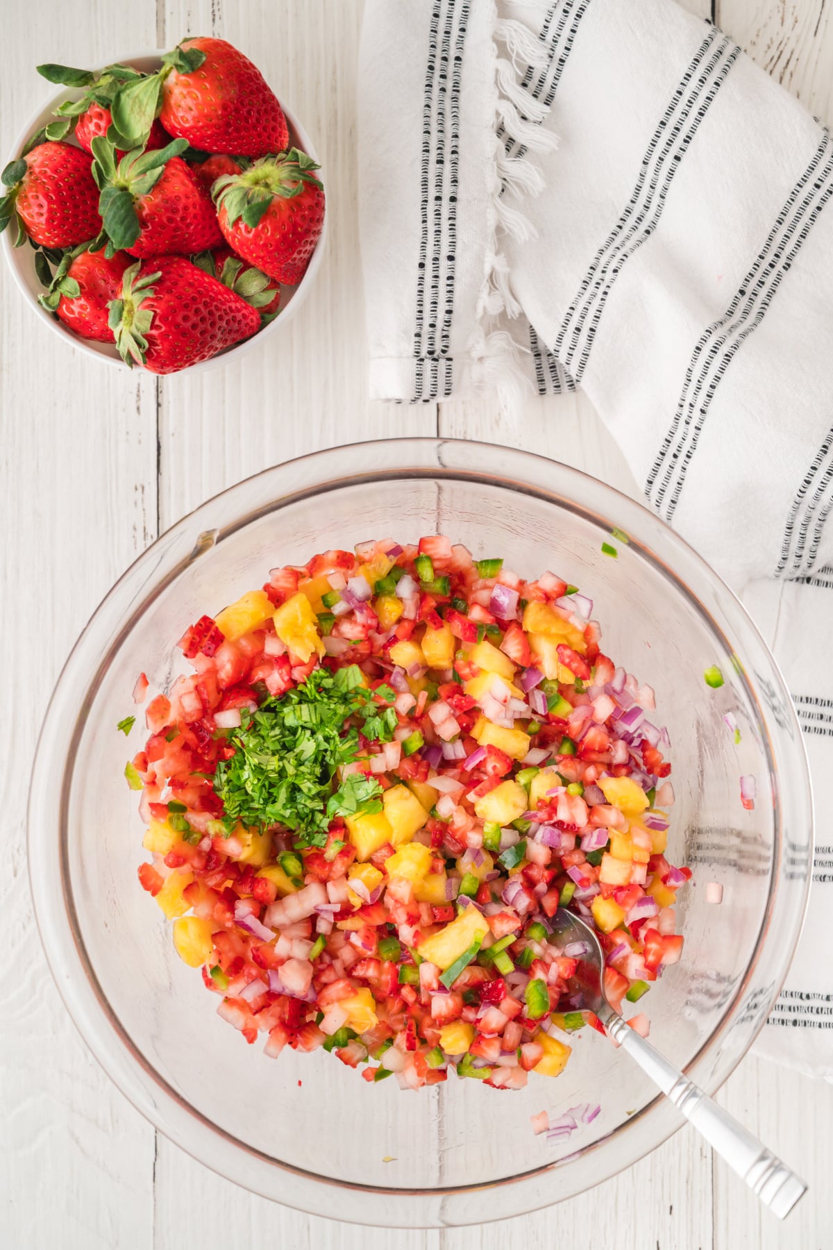 A glass bowl filled with chopped strawberries, pineapple, onion, and cilantro, next to a bowl of whole strawberries and a white striped kitchen towel on a white wooden surface.