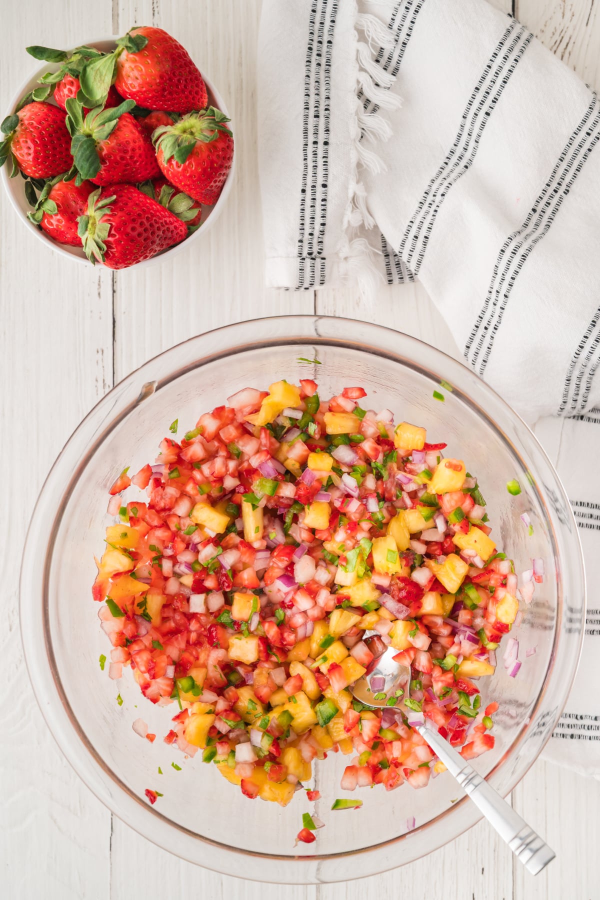 A glass bowl of strawberry salsa with diced fruit, onions, and cilantro sits next to a bowl of whole strawberries and a striped kitchen towel on a white wooden surface.