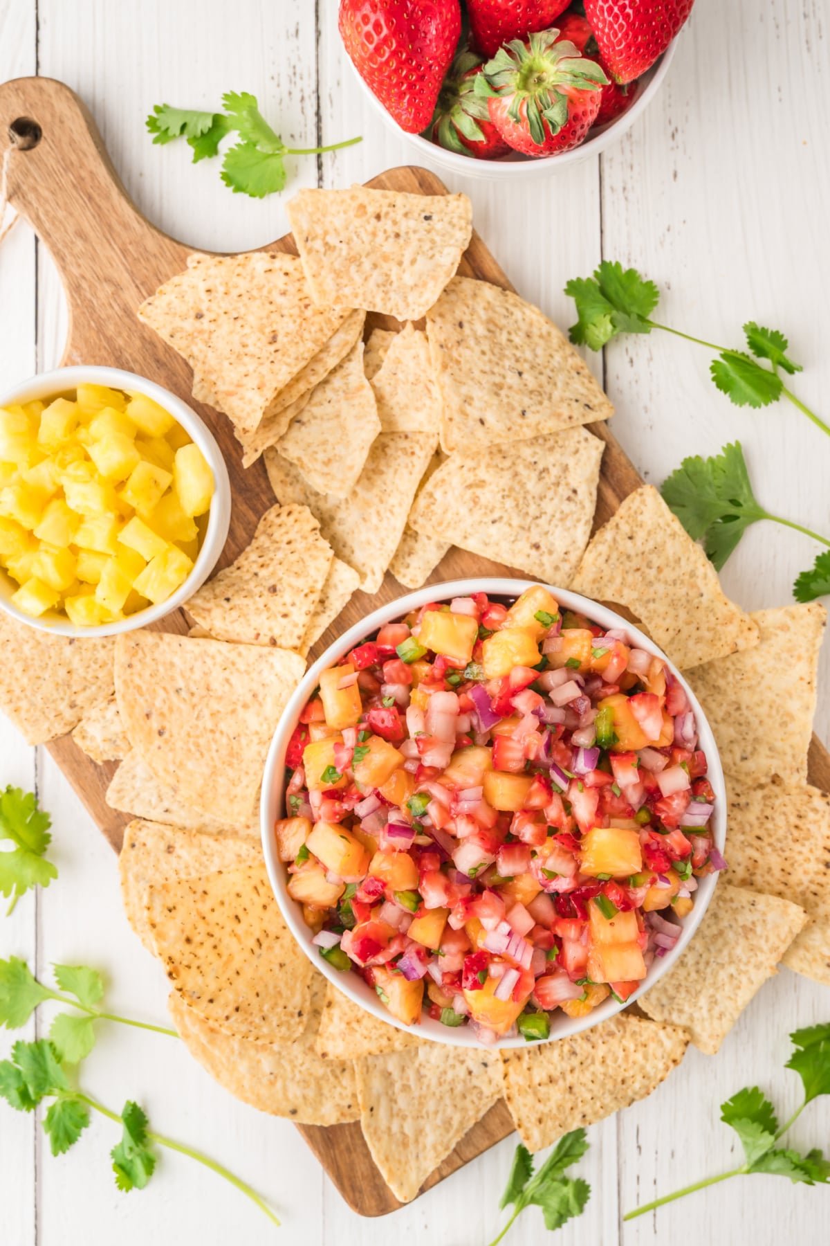 A bowl of fruit salsa surrounded by tortilla chips on a wooden board, with a small bowl of diced pineapple and a bowl of fresh strawberries nearby.