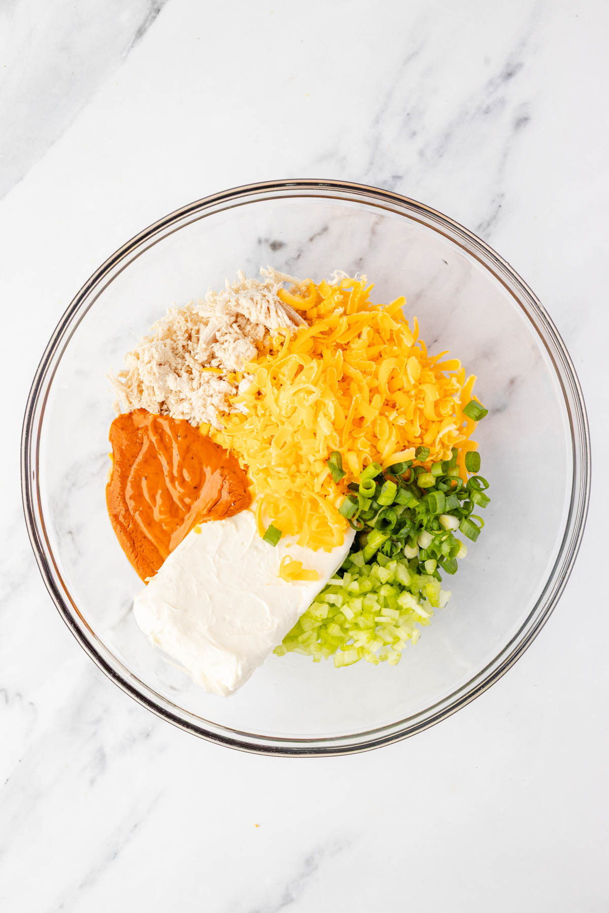 A glass bowl containing shredded chicken, shredded cheddar cheese, sliced green onions, chopped celery, buffalo sauce, and cream cheese on a marble surface.