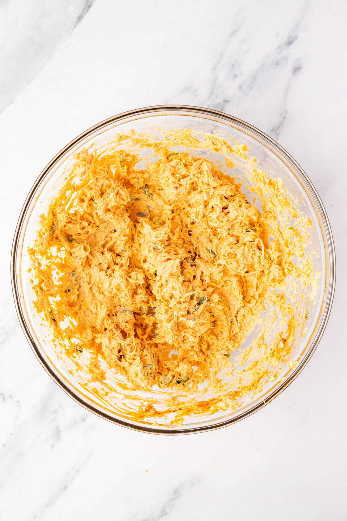 A glass bowl filled with a mixture of shredded cheese, spices, and chopped ingredients on a white marble surface.