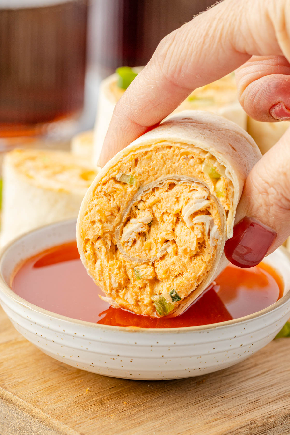 A hand dips a rolled tortilla filled with a creamy orange chicken mixture into a bowl of red sauce.