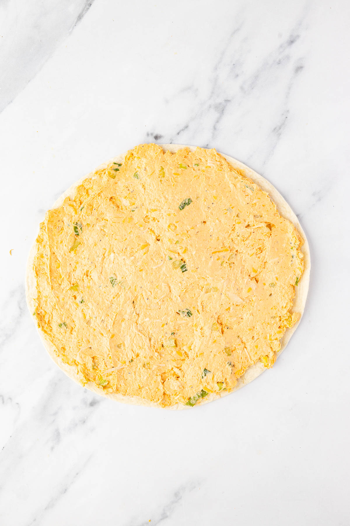 A round, flat piece of dough topped with an even layer of orange cheese and green onion mixture on a white marble surface.