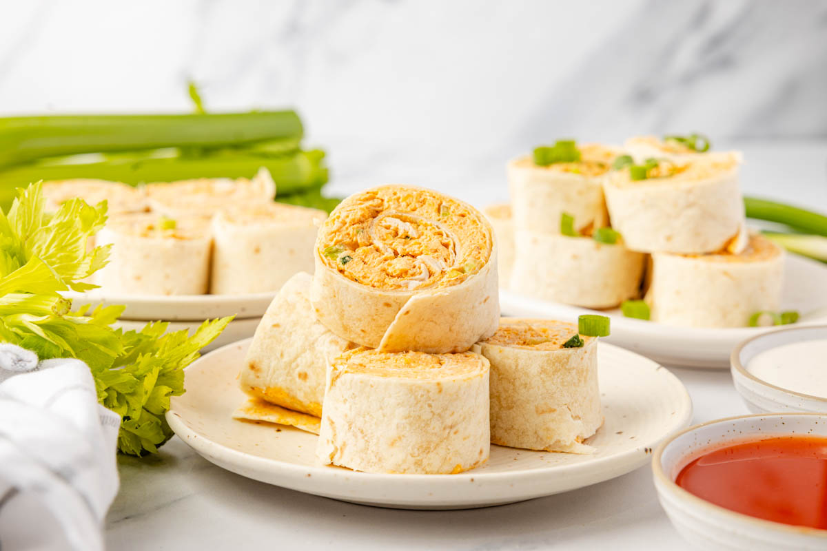A plate of tortilla pinwheels filled with a creamy orange mixture, garnished with green onions, with celery, dipping sauces, and more pinwheels in the background.