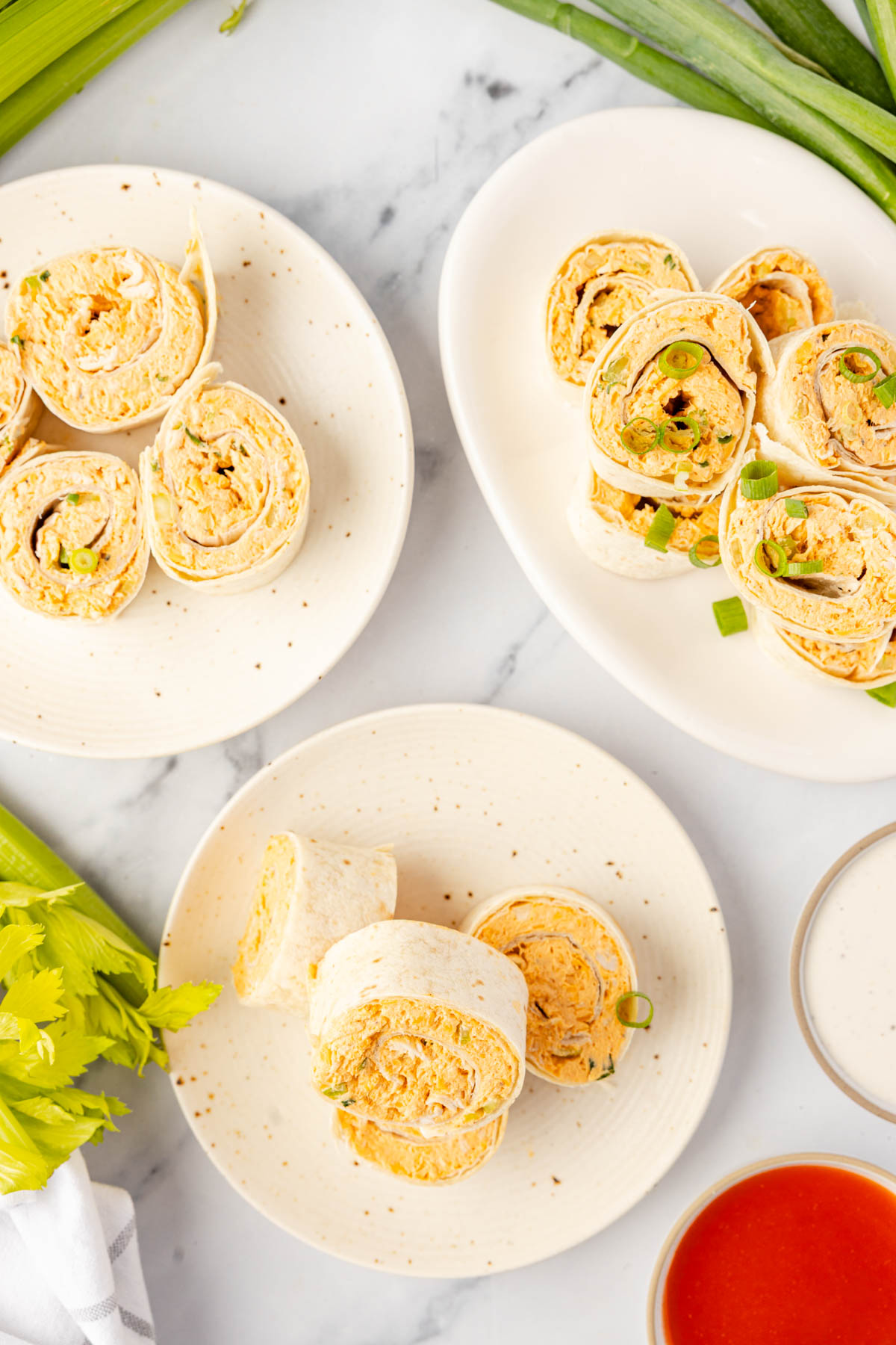 Three plates with sliced tortilla pinwheels filled with a creamy, orange-tinted mixture, garnished with green onions; celery, green onions, and dipping sauces on the side.