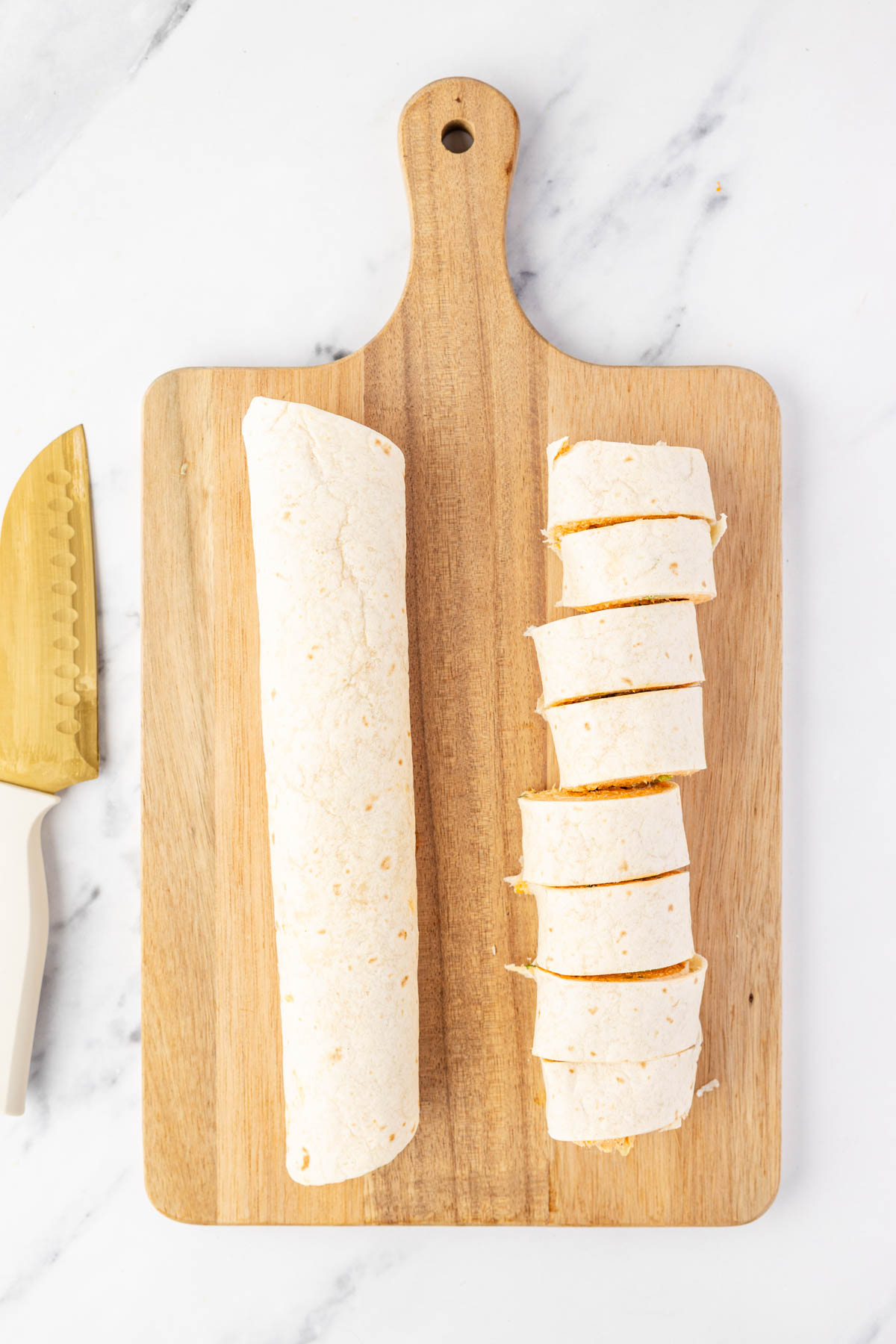 A whole tortilla roll and several sliced pieces are arranged on a wooden cutting board next to a serrated knife on a white surface.